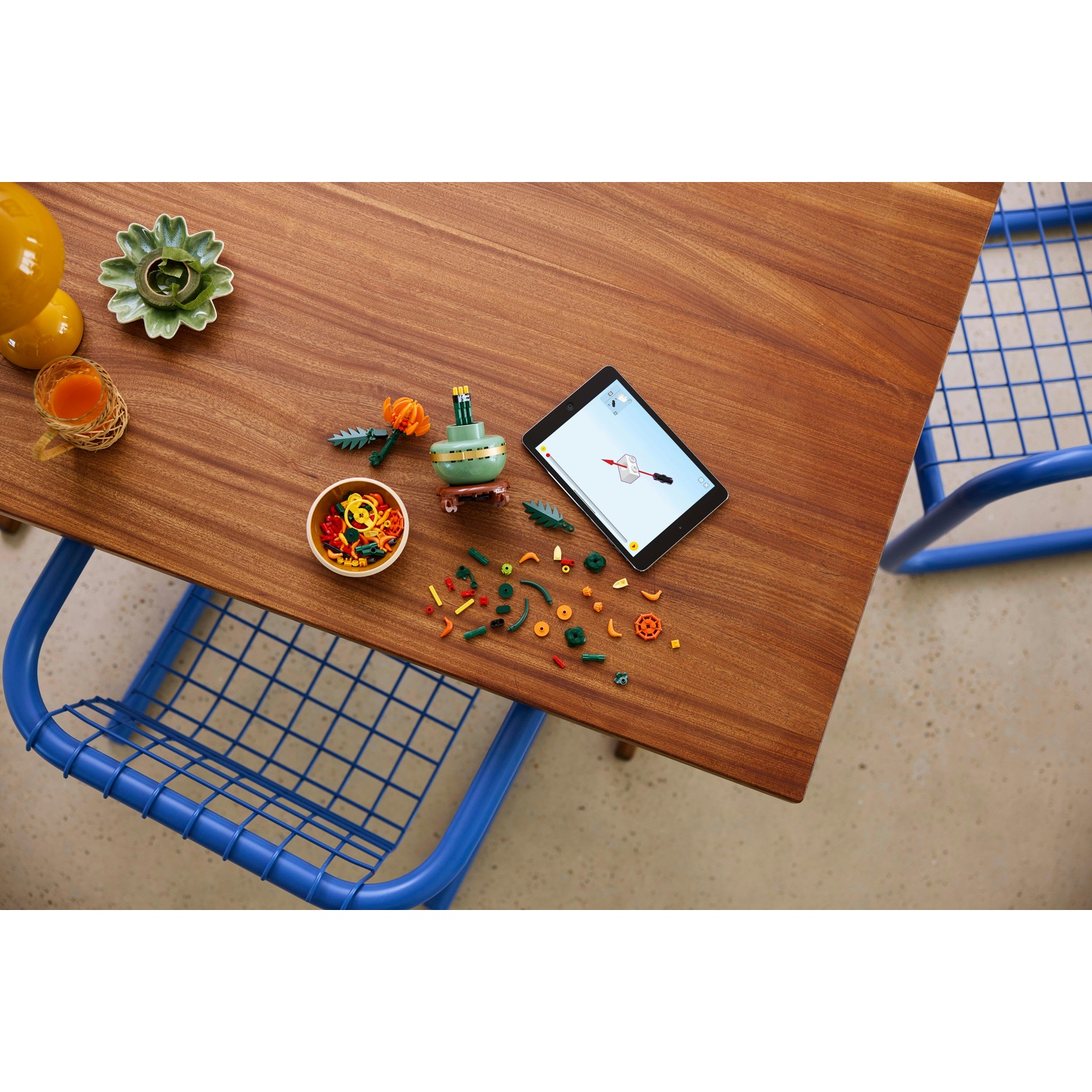 Un tavolo di legno con tablet, mattoncini e una piccola pianta. Sedia blu a destra. Bevande e piatto decorativo sul tavolo.