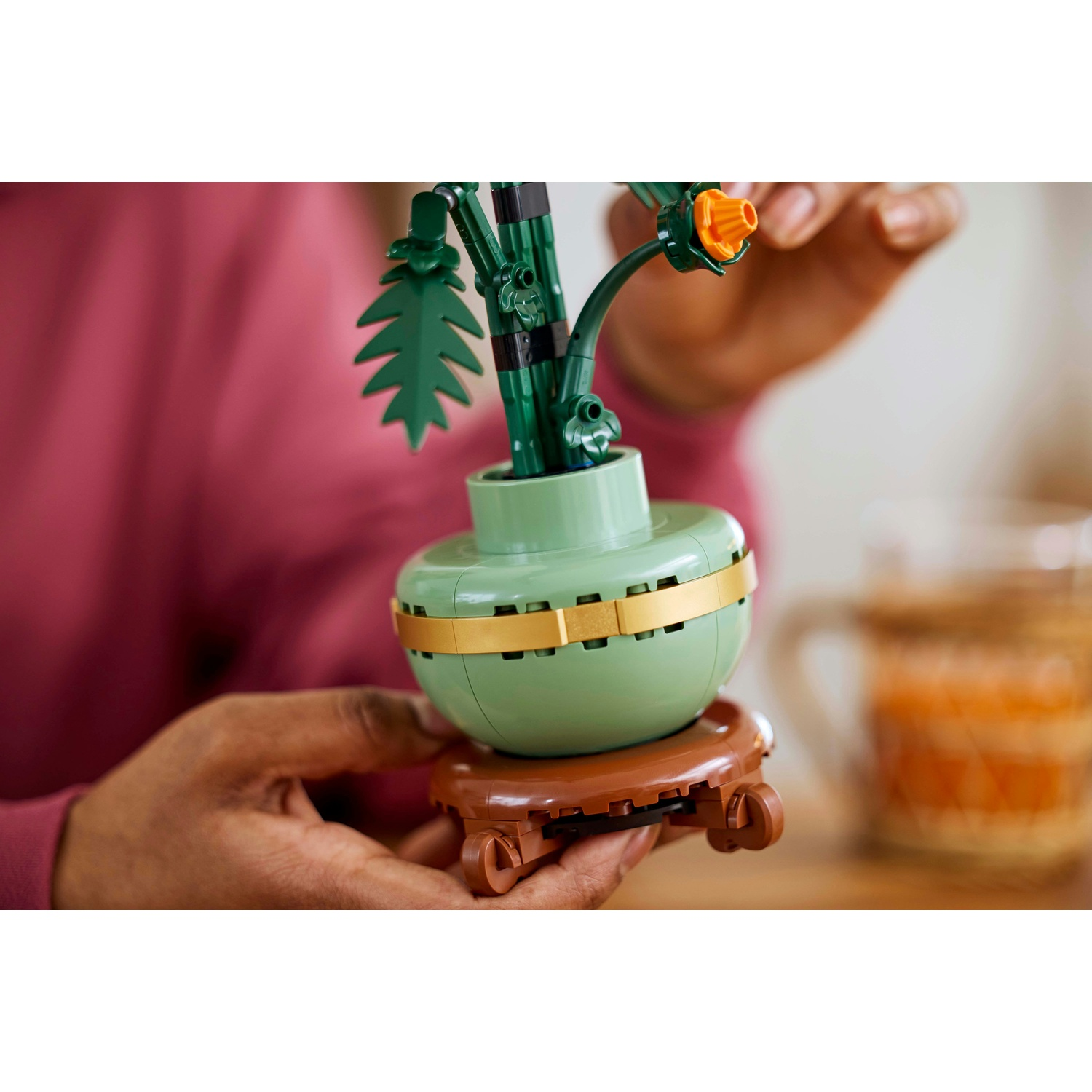 Una persona tiene in mano una pianta Lego. La pianta ha steli e foglie verdi e un vaso verde chiaro. Il vaso poggia su un supporto marrone. Una mano è sullo sfondo.