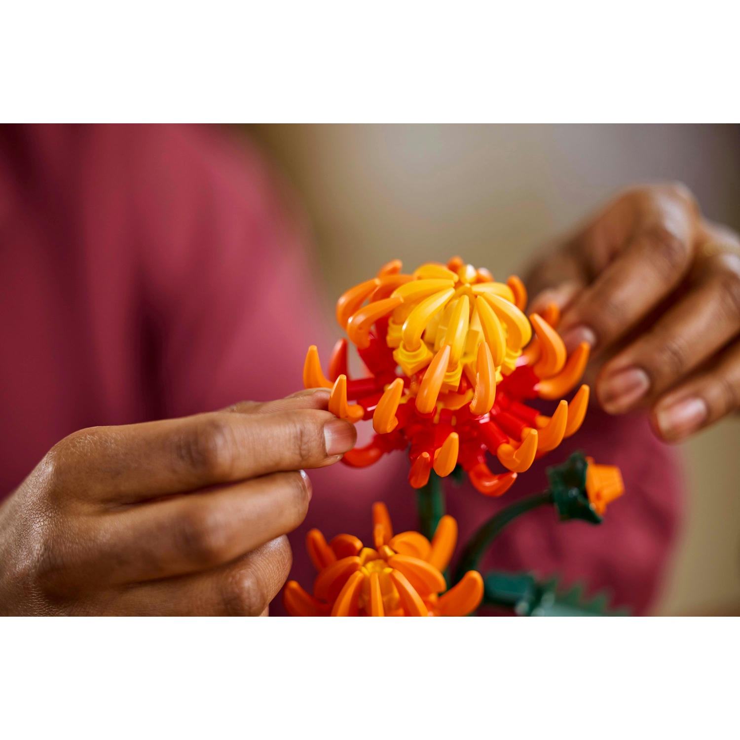 Mani che tengono un fiore di plastica colorato. Il fiore è arancione e giallo, con steli verdi. Una persona indossa una camicia rosa.