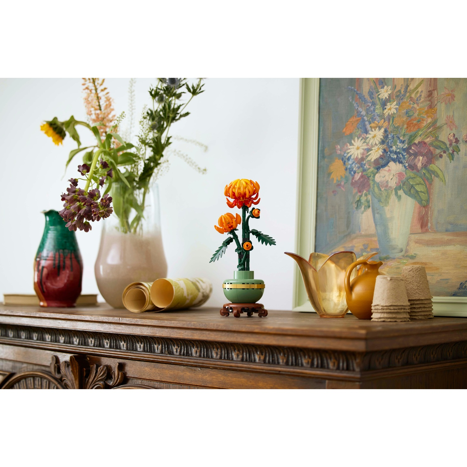 Tavolo in legno con vasi di fiori, un dipinto e un fiore Lego. Colori: arancione, verde, marrone e bianco.