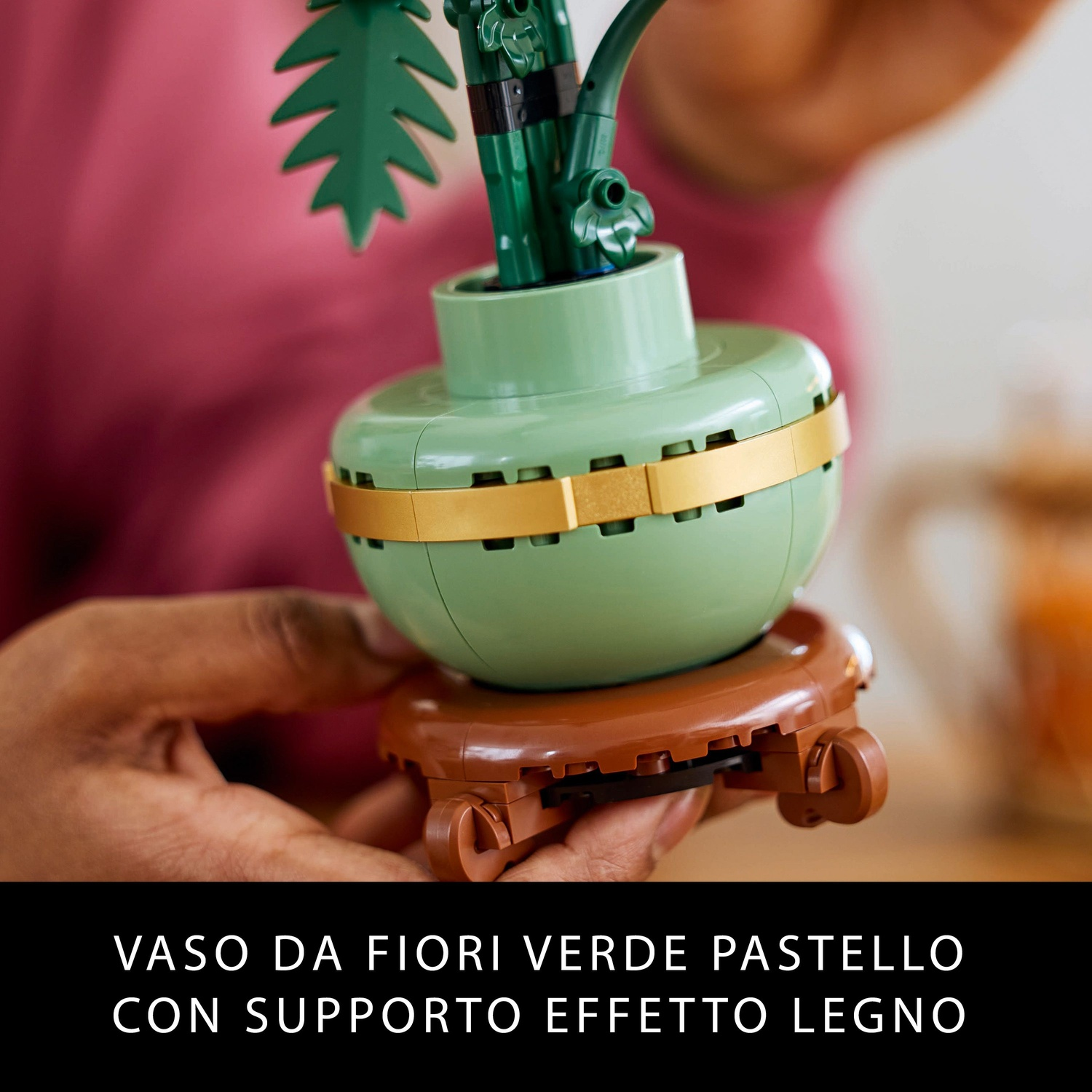 Un vaso di fiori verde pastello con una fascia dorata e un supporto effetto legno marrone, che contiene una pianta verde. Una persona lo tiene.
