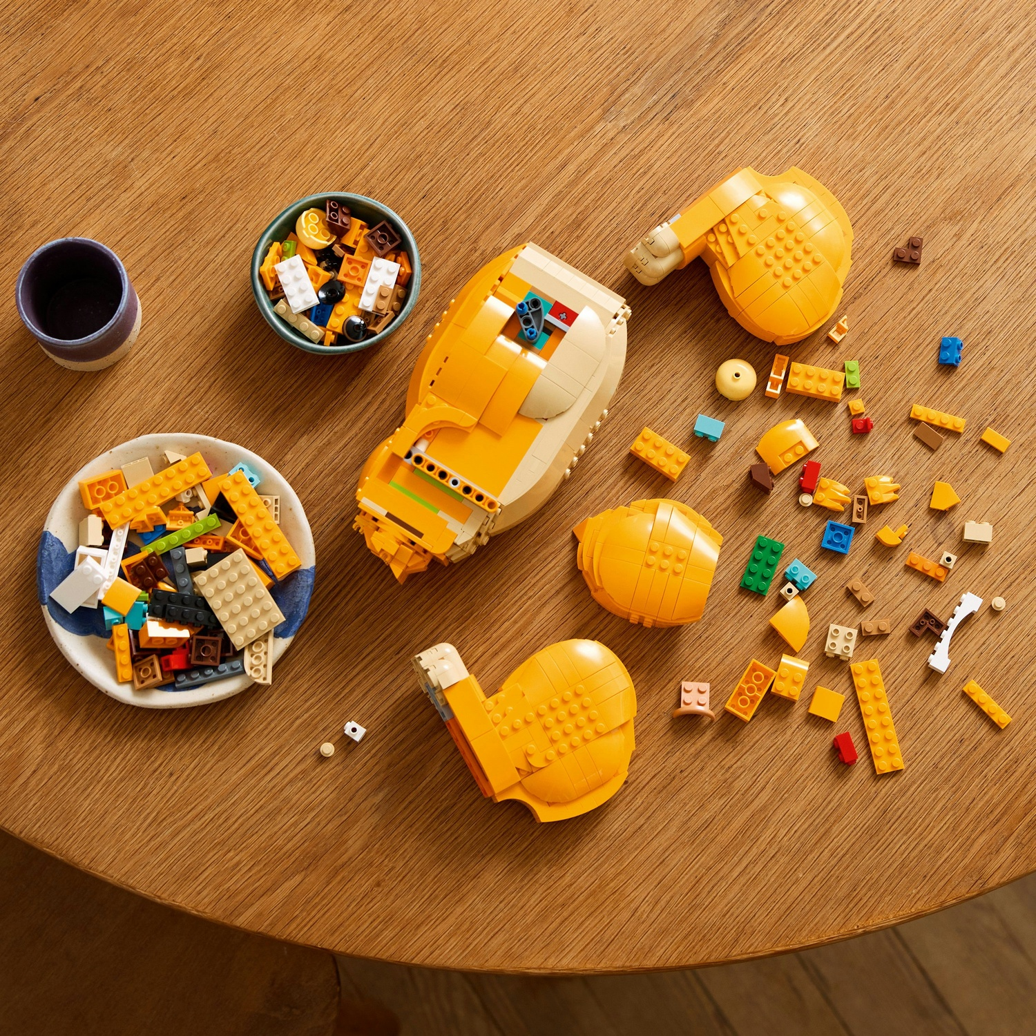 Tavolo di legno con mattoncini Lego, ciotole di mattoncini e una tazza viola. È presente una struttura Lego gialla parzialmente assemblata.