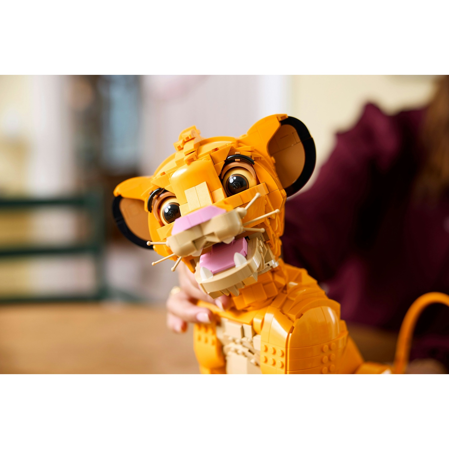 Un giocattolo Lego di Simba è tenuto in mano da una persona. Il giocattolo è arancione e beige con occhi neri. Lo sfondo è sfocato.