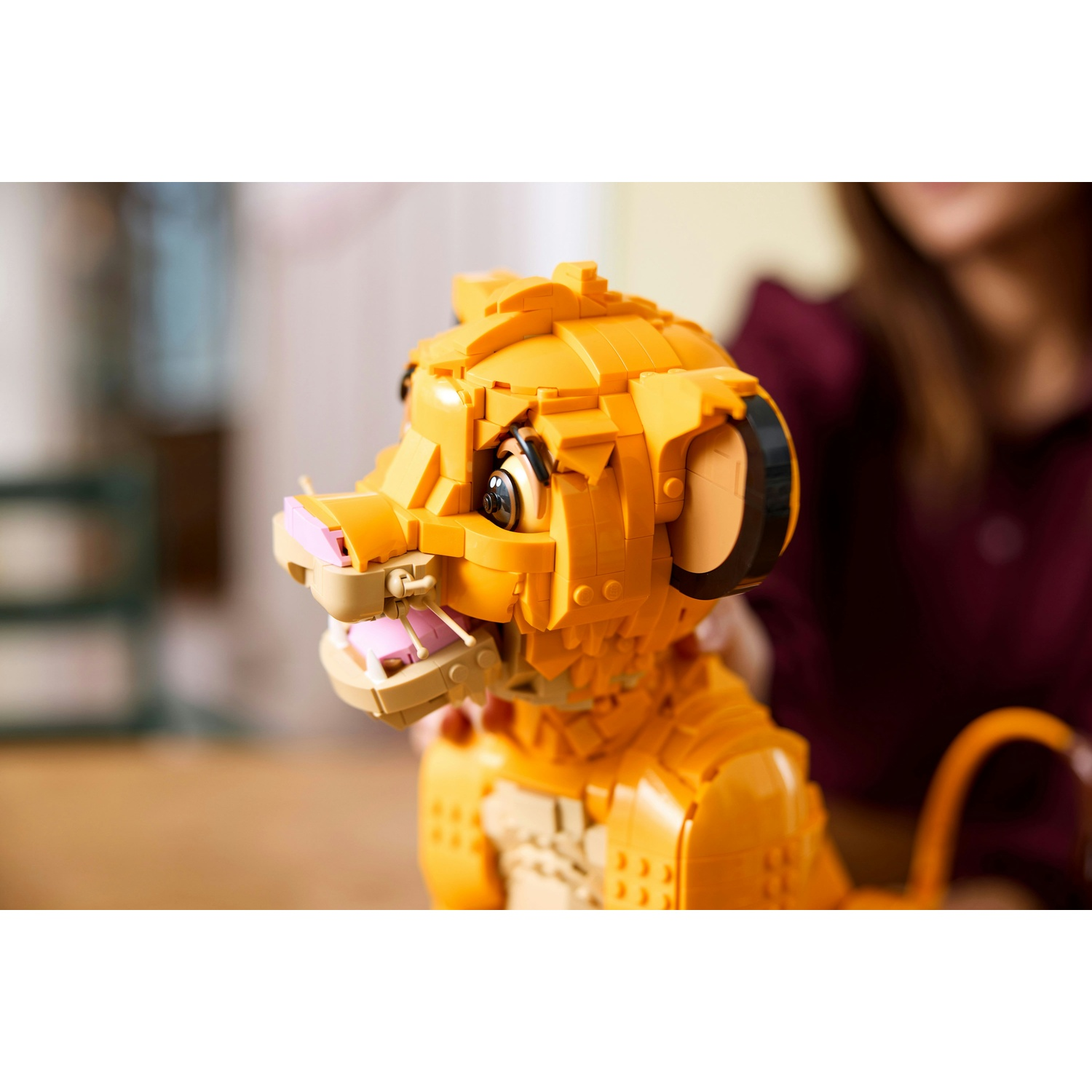 Un modello Lego Simba, in giallo e beige, è esposto. Il volto del cucciolo di leone è a fuoco, con bocca rosa e mascelle aperte. Una persona sullo sfondo.