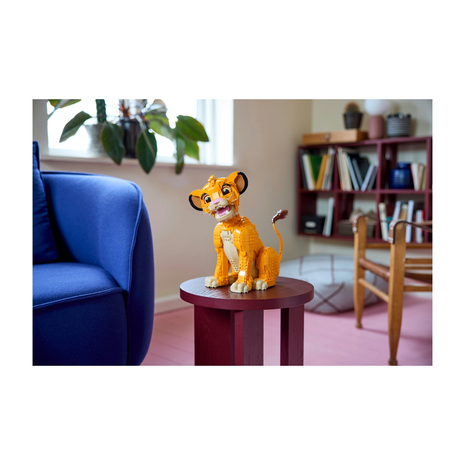 Una figura Lego di Simba è seduta su un tavolo rosso. A sinistra c'è una sedia blu, sullo sfondo una libreria e una sedia.