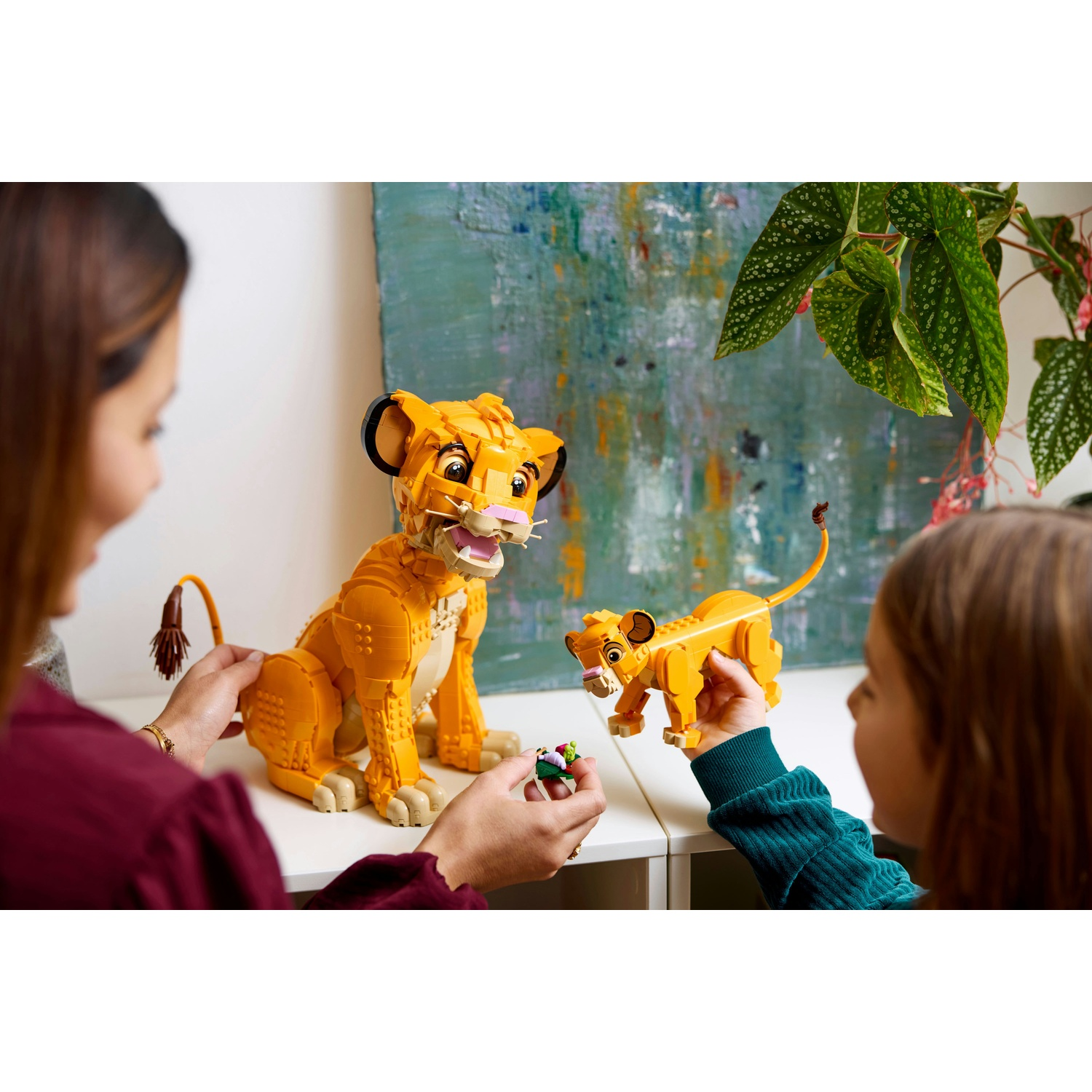 Due persone giocano con i personaggi Lego Simba. Una grande figura è su un tavolo. Un bambino tiene in mano un Simba più piccolo. Lo sfondo ha un dipinto e delle piante.