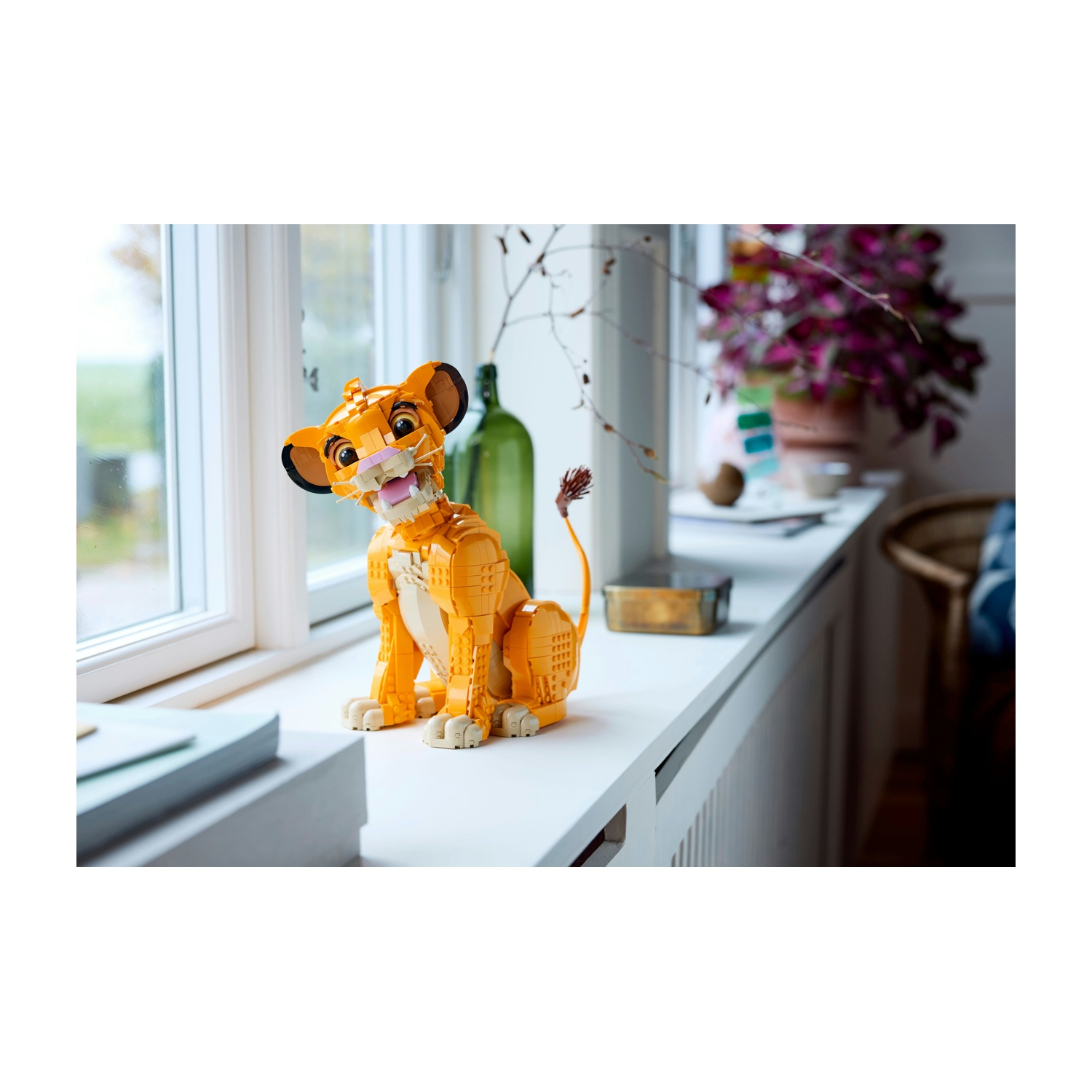 Un modello Lego di Simba è seduto su un davanzale. È arancione con la bocca rosa. Dietro c'è una bottiglia verde e fiori viola.