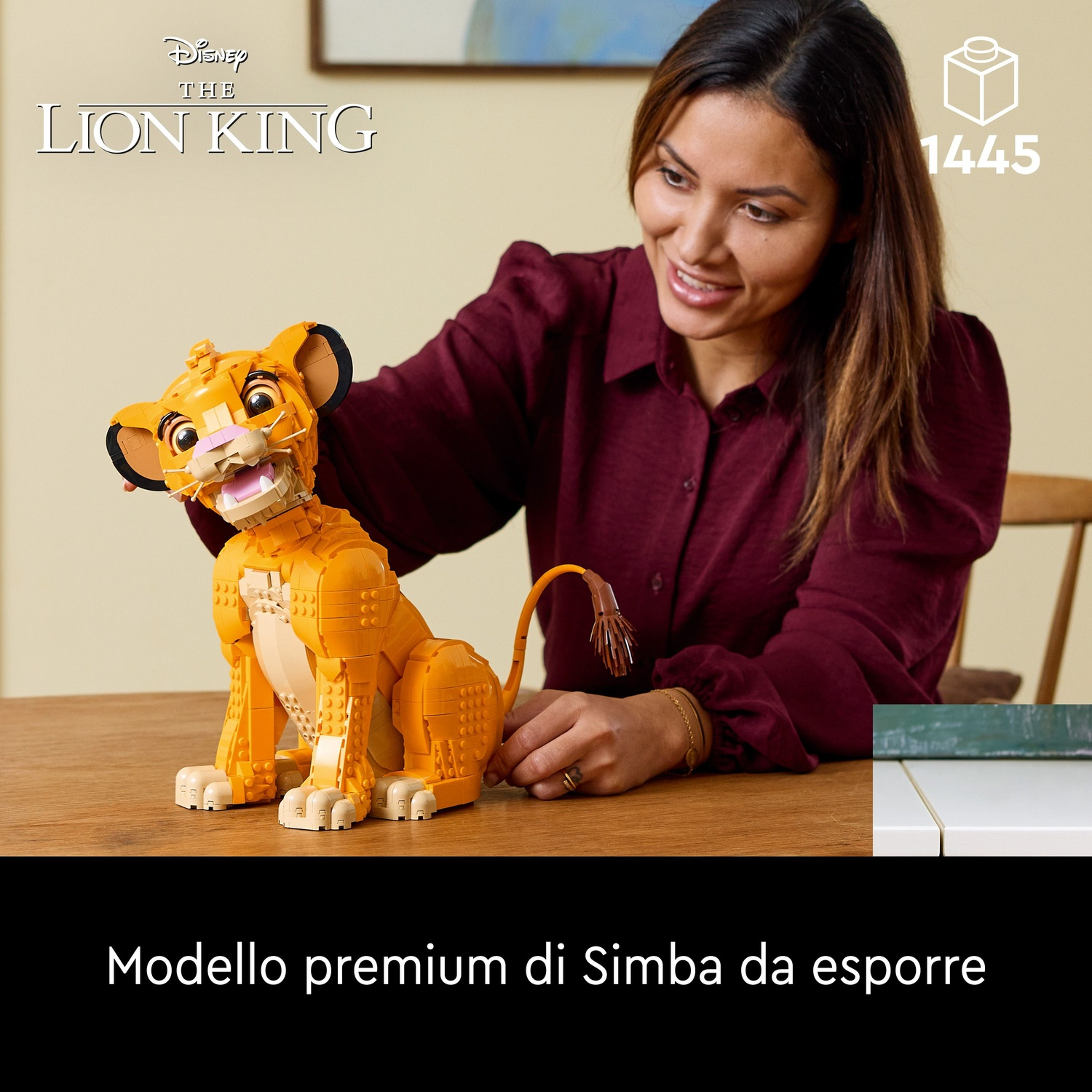 Una donna sorride, tenendo un Simba Lego. Il Simba Lego è arancione e bianco. Lo sfondo include testo e un tavolo.