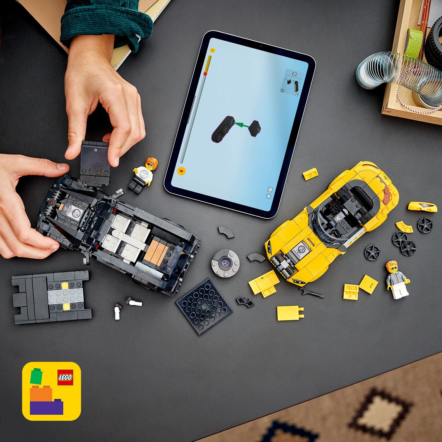 Mani che assemblano un'auto Lego nera, accanto a un'auto Lego gialla. Un tablet mostra le istruzioni. Pezzi e figure Lego sparsi.