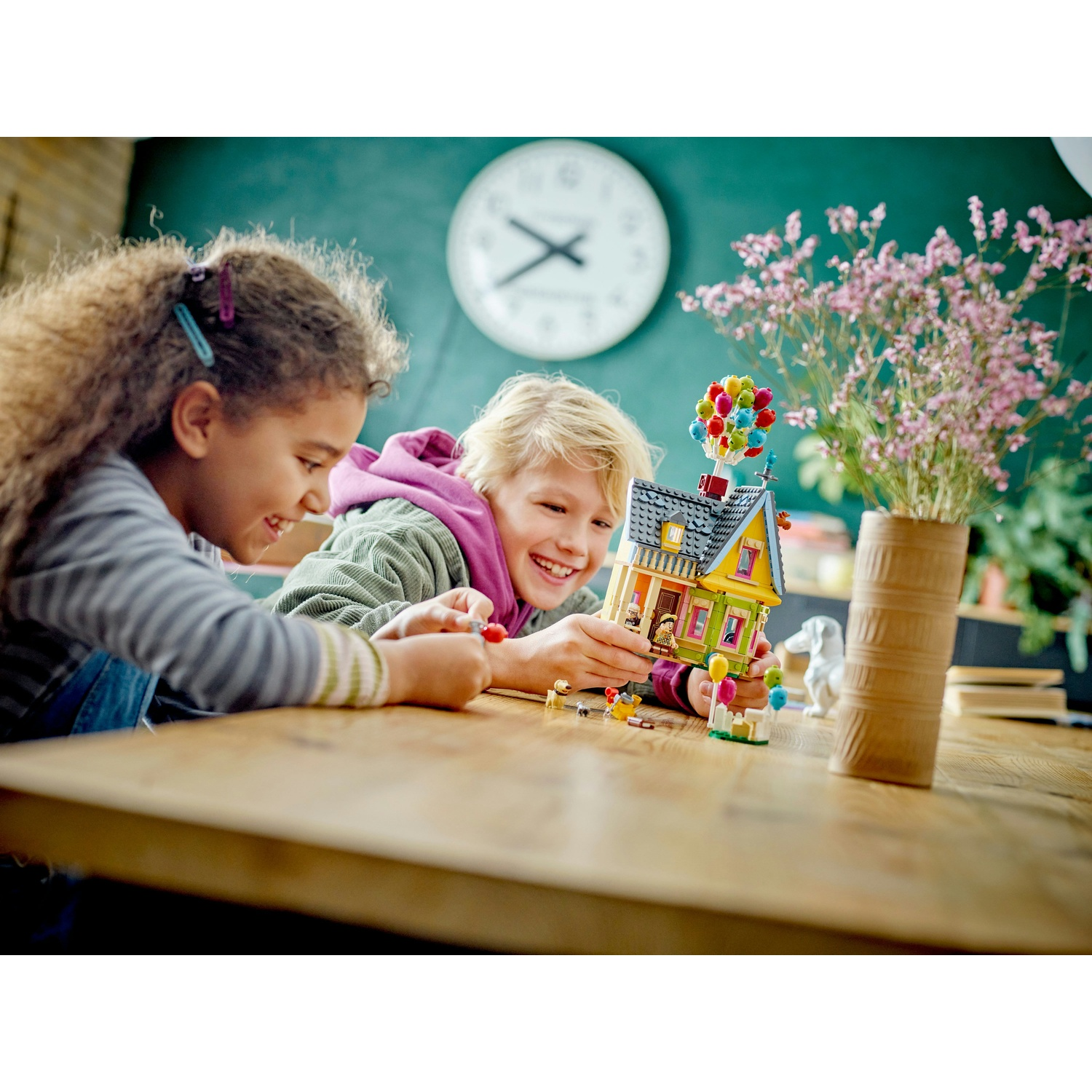 Due bambini sorridono mentre giocano con una casa Lego. Un orologio è appeso al muro e dei fiori sono in un vaso.