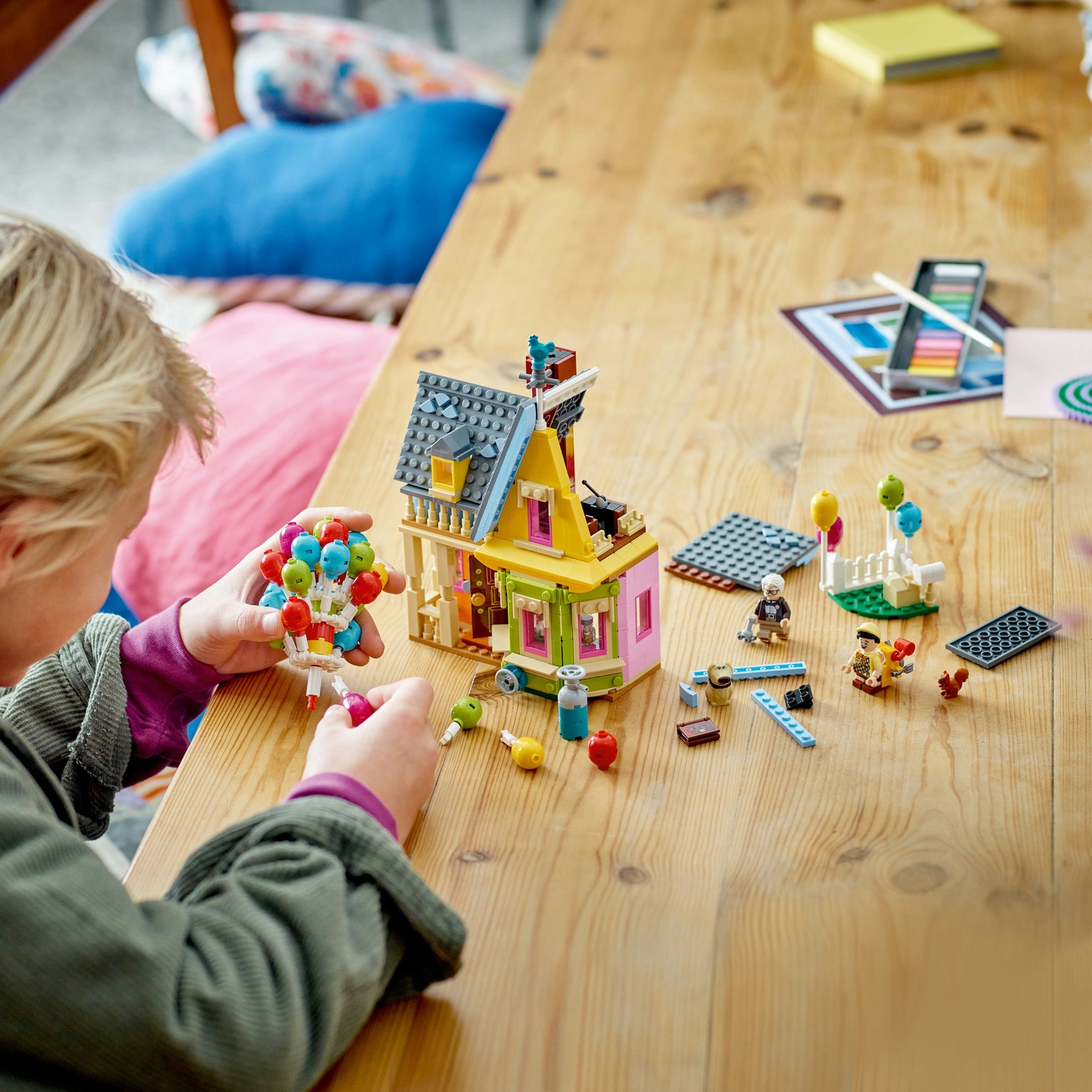 Un bambino sta costruendo una casa Lego con palloncini. La scena include personaggi Lego, una casa e vari accessori Lego su un tavolo di legno.