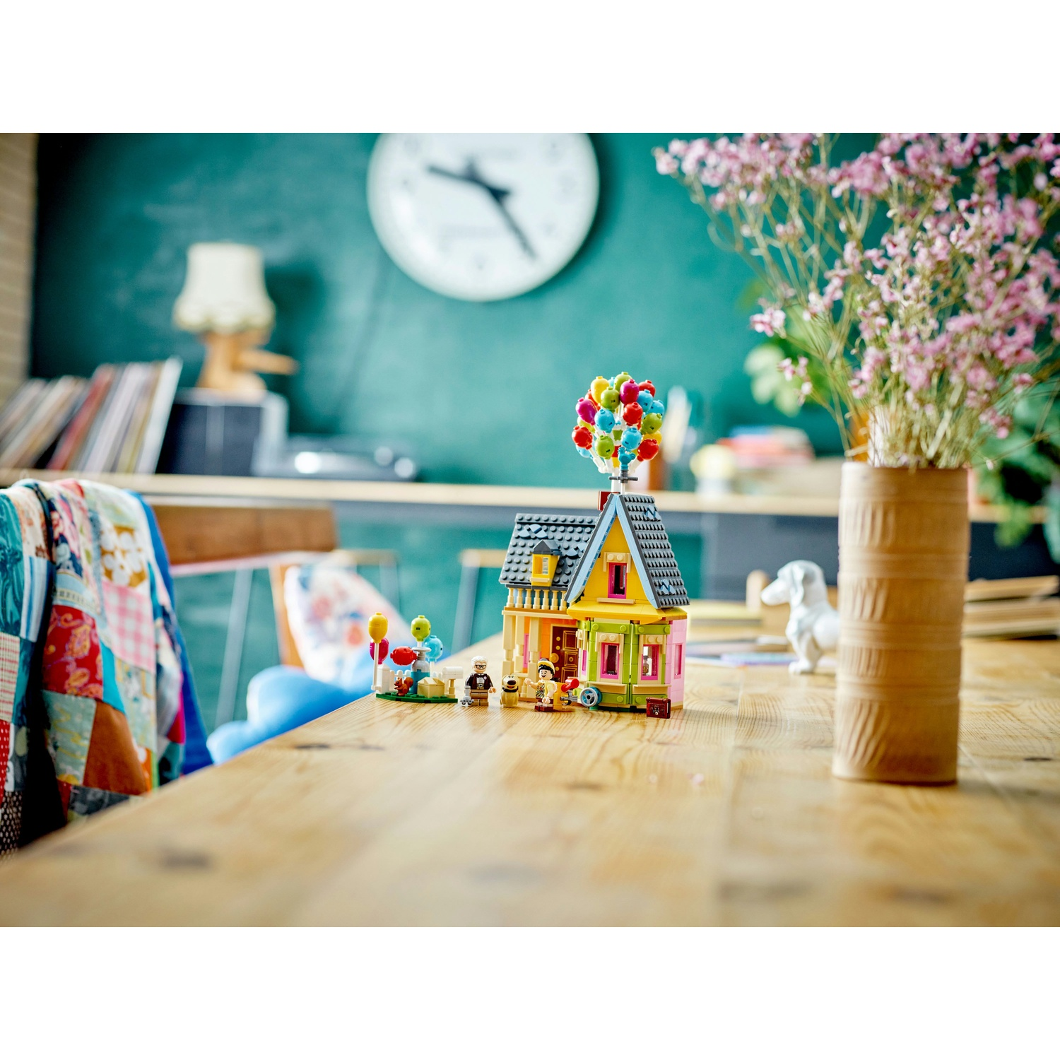 Una casa Lego con palloncini, statuine e un vaso di fiori su un tavolo di legno. Un orologio e una lampada sullo sfondo.