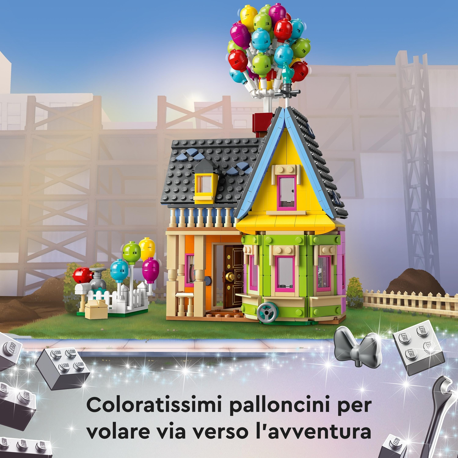 Una casa Lego con palloncini colorati attaccati. La casa è su una base verde, circondata da una recinzione bianca. Lo sfondo è un cantiere.