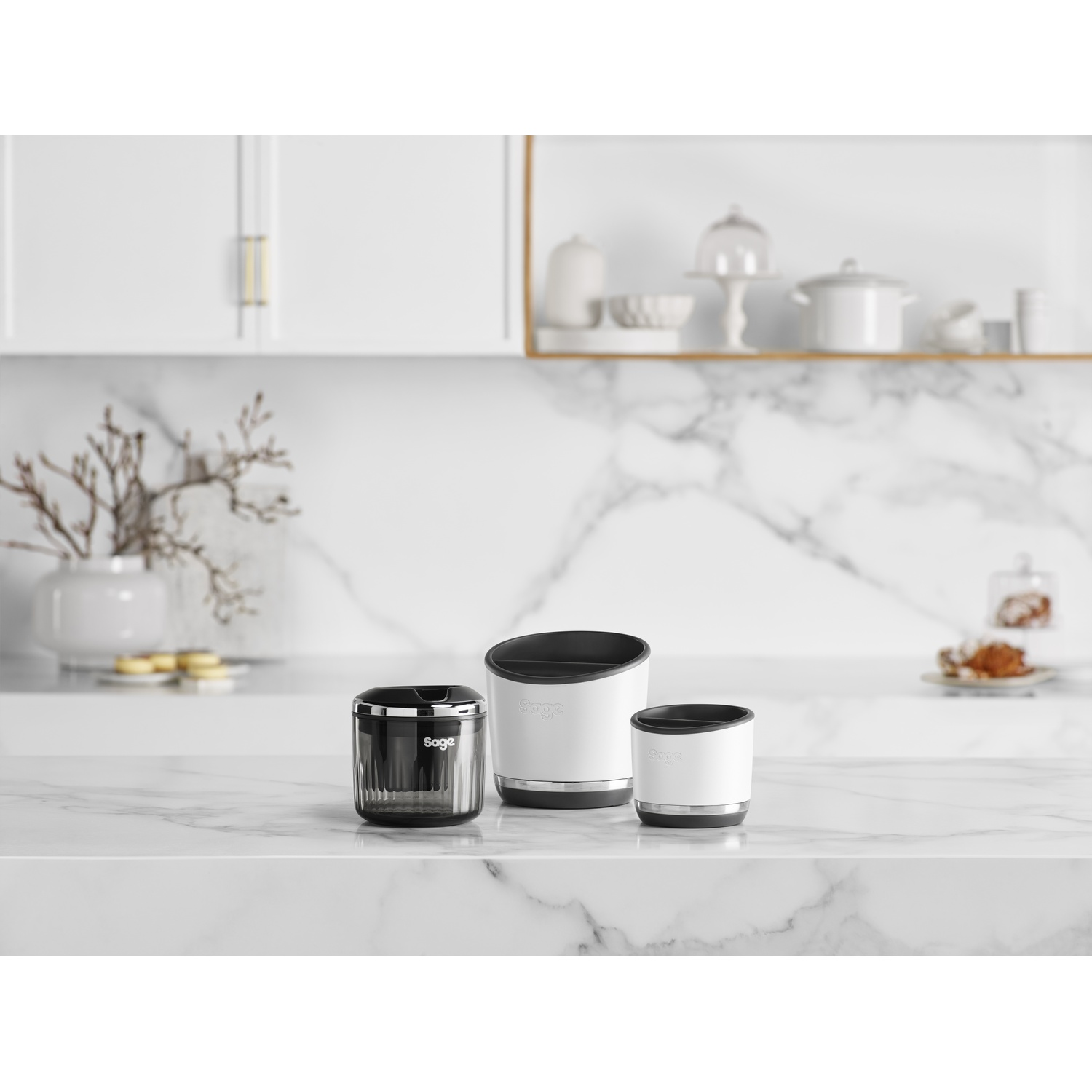Three white kitchen containers with black tops on a marble surface. Background has white cabinets and shelves with kitchenware.