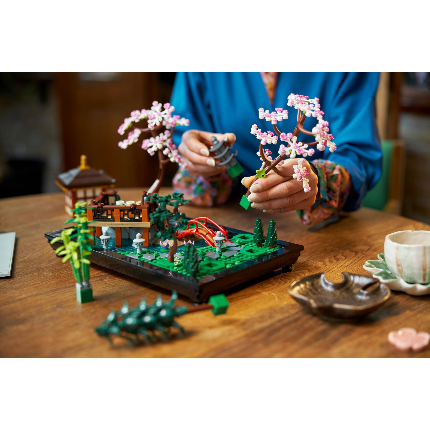 Una persona che assembla un bonsai Lego. La scena comprende un giardino Lego con un ponte rosso, una casa e altri dettagli.