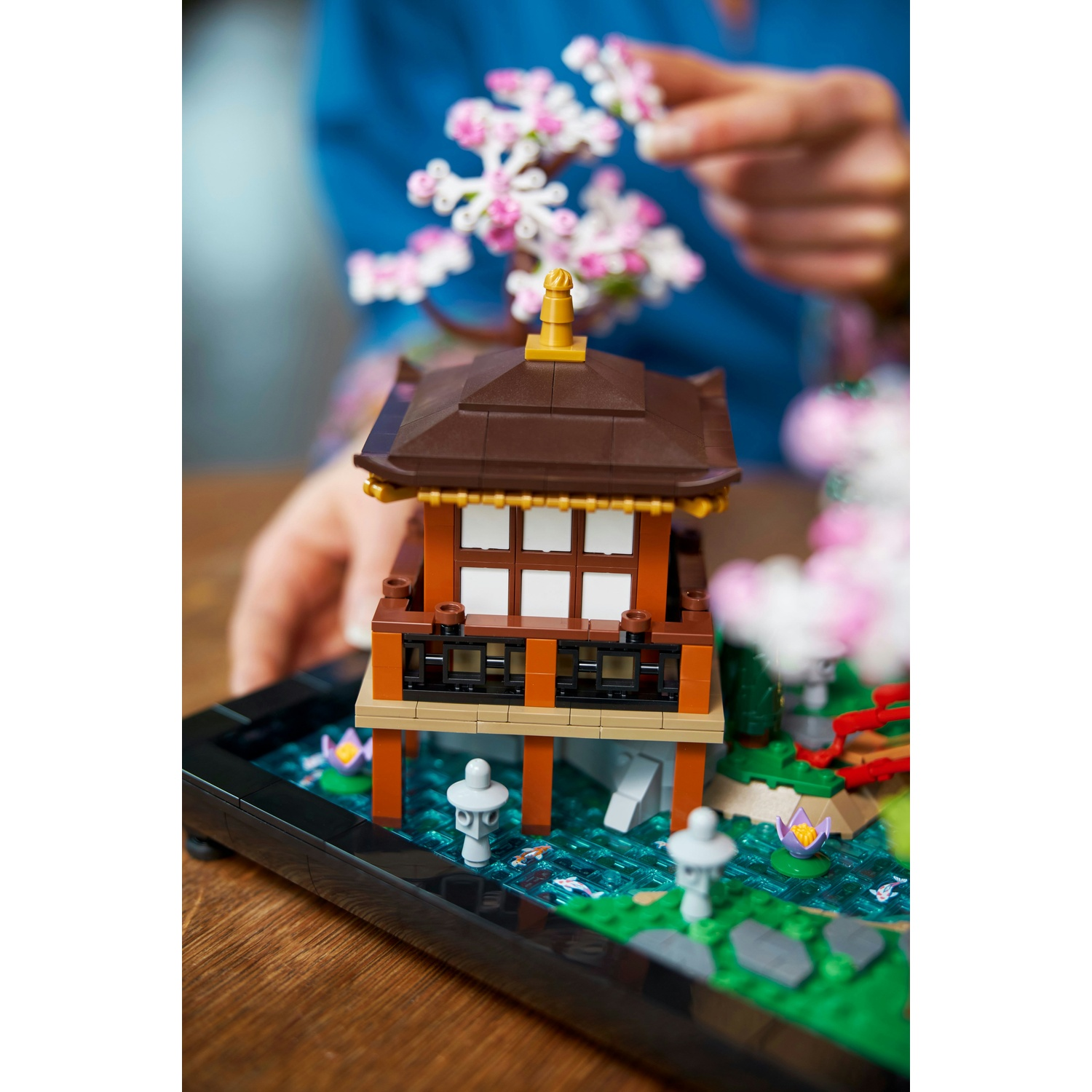 Un tempio giapponese Lego con fiori di ciliegio. Una persona tiene i fiori. Il tempio si trova sopra uno stagno. La scena è su un tavolo di legno.