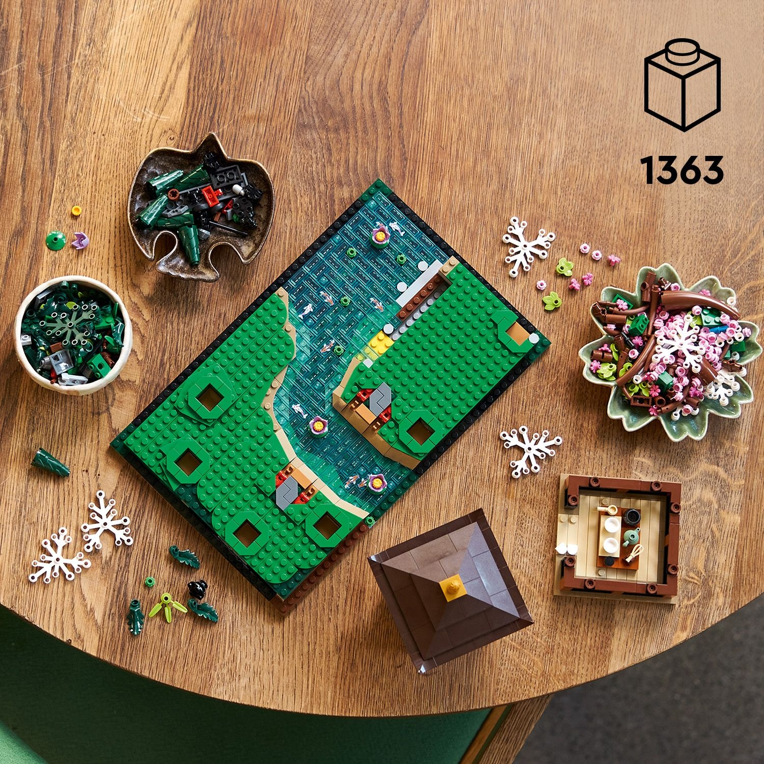 Vista dall'alto di un set Lego. Una baseplate verde con un fiume, edifici e pezzi circostanti come fiori e alberi su legno.