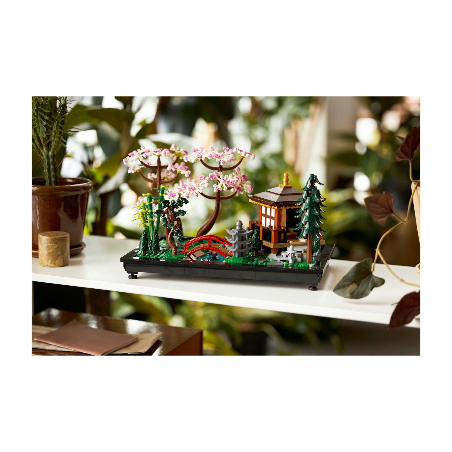 Un modello Lego di giardino giapponese con una pagoda, un ponte, alberi e bambù. È su una mensola bianca con piante sullo sfondo.