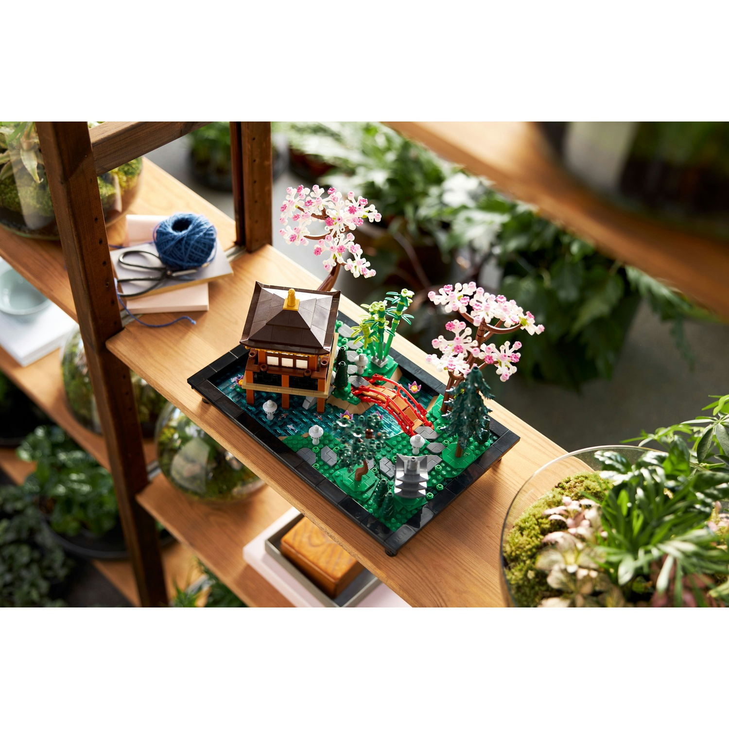 Una scena Lego di giardino giapponese su una mensola di legno, con un ponte rosso, una pagoda e alberi in fiore, circondata da piante.