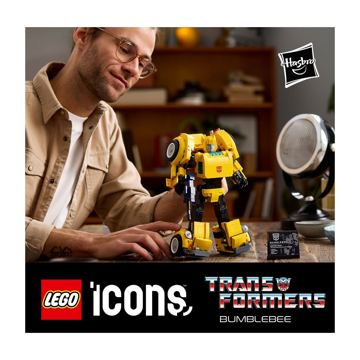 Un uomo tiene in mano un personaggio Lego di Bumblebee giallo. Sono visibili i loghi Hasbro e Lego. Lo sfondo mostra mensole di legno e una lampada.