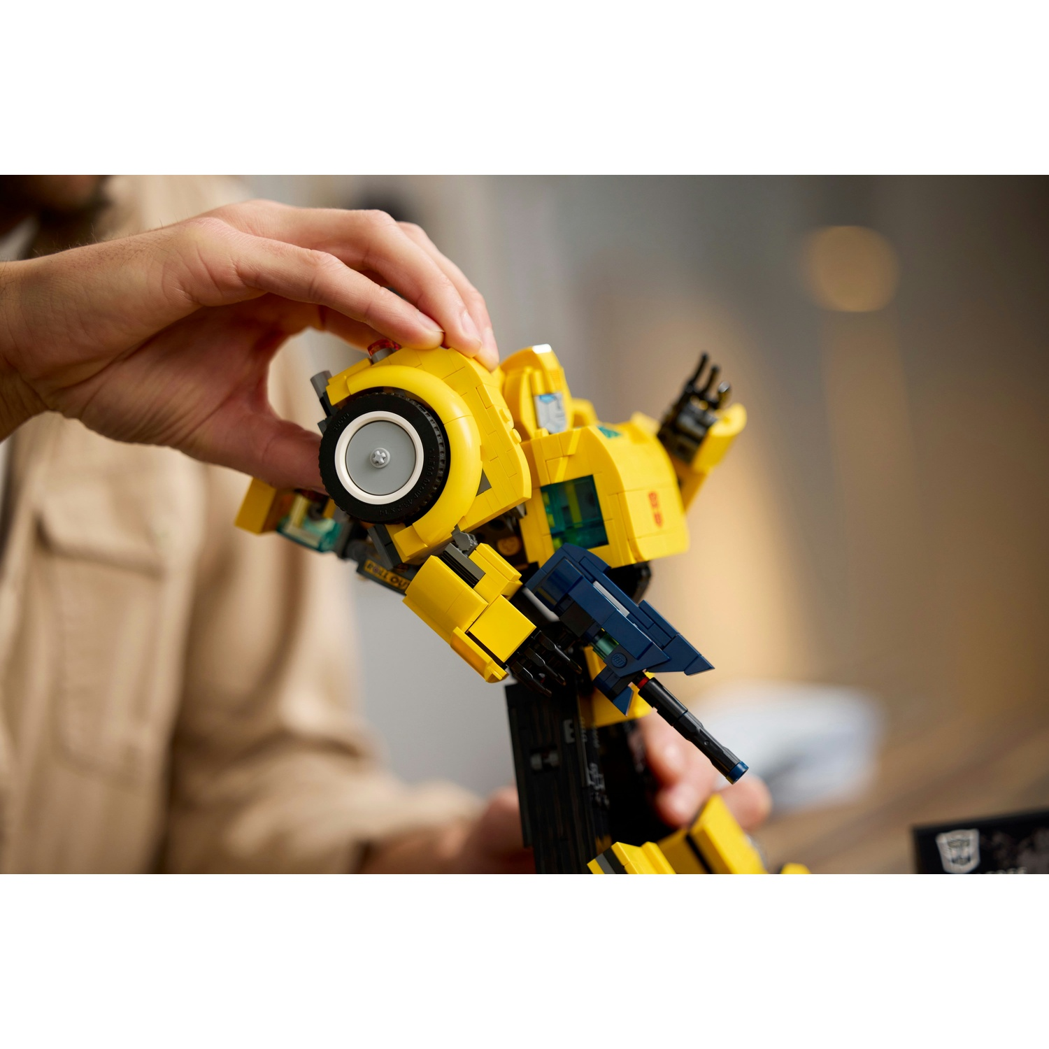 Una persona tiene in mano un Transformer Lego giallo. Il giocattolo è Bumblebee, con dettagli neri e blu. Le mani tengono il giocattolo, davanti a uno sfondo sfocato.