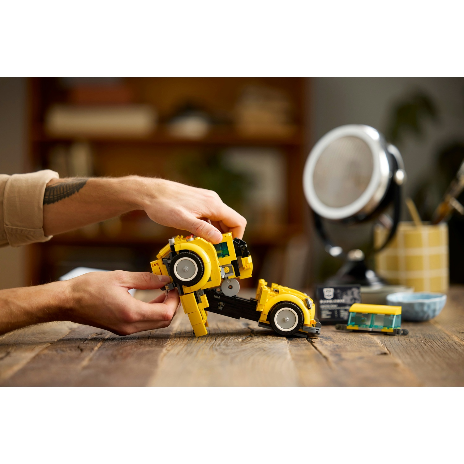 Mani che assemblano un'auto Lego gialla su un tavolo di legno. Altri pezzi Lego e una lampada decorativa sullo sfondo.