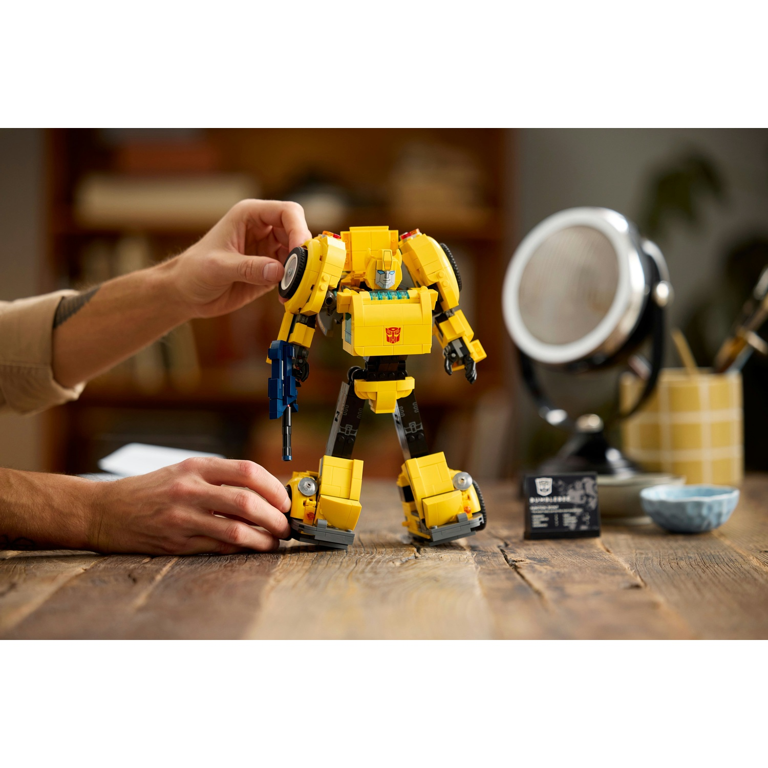 Un giocattolo Lego Bumblebee giallo è su un tavolo di legno. Le mani di una persona regolano il giocattolo. Un microfono e una carta sullo sfondo.