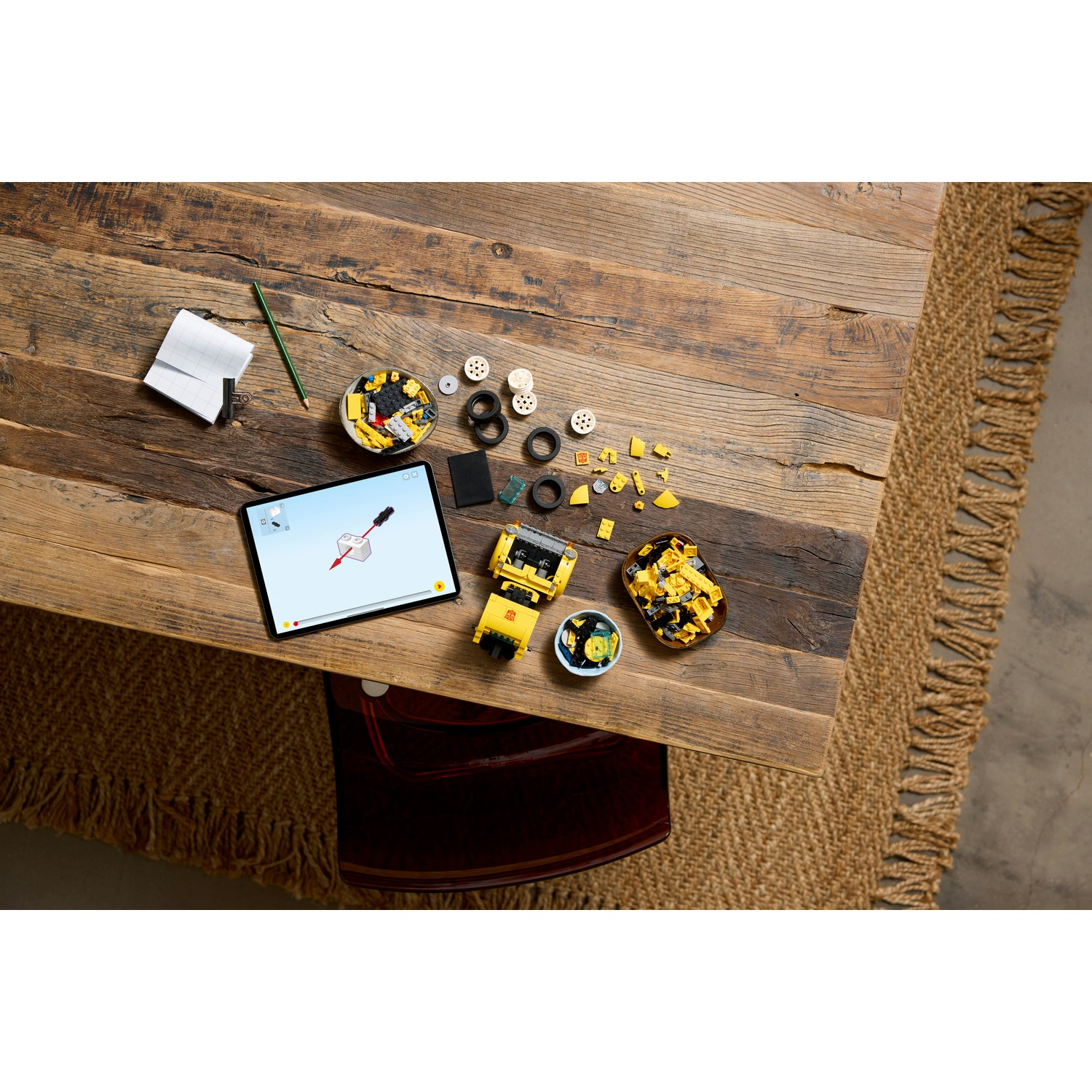 Tavolo di legno con pezzi Lego, tablet e matita. Un'auto Lego è al centro. Pezzi sparsi.