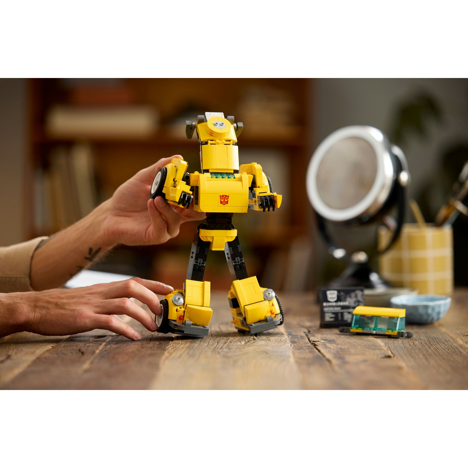 Una figura Lego di Bumblebee gialla è su un tavolo di legno. Le mani di una persona tengono la figura. Lo sfondo include uno specchio e dei libri.