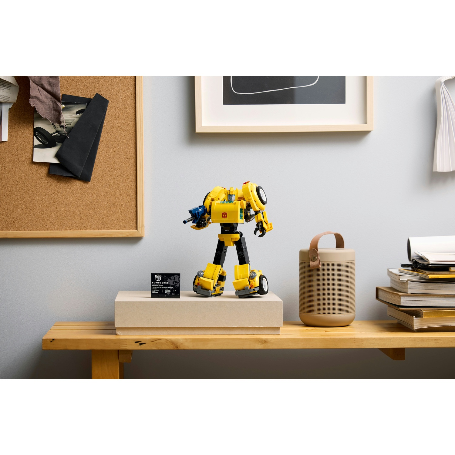 Un giocattolo Bumblebee giallo e nero è su una mensola di legno con un altoparlante, libri e una bacheca. Un'arte incorniciata è sul muro.