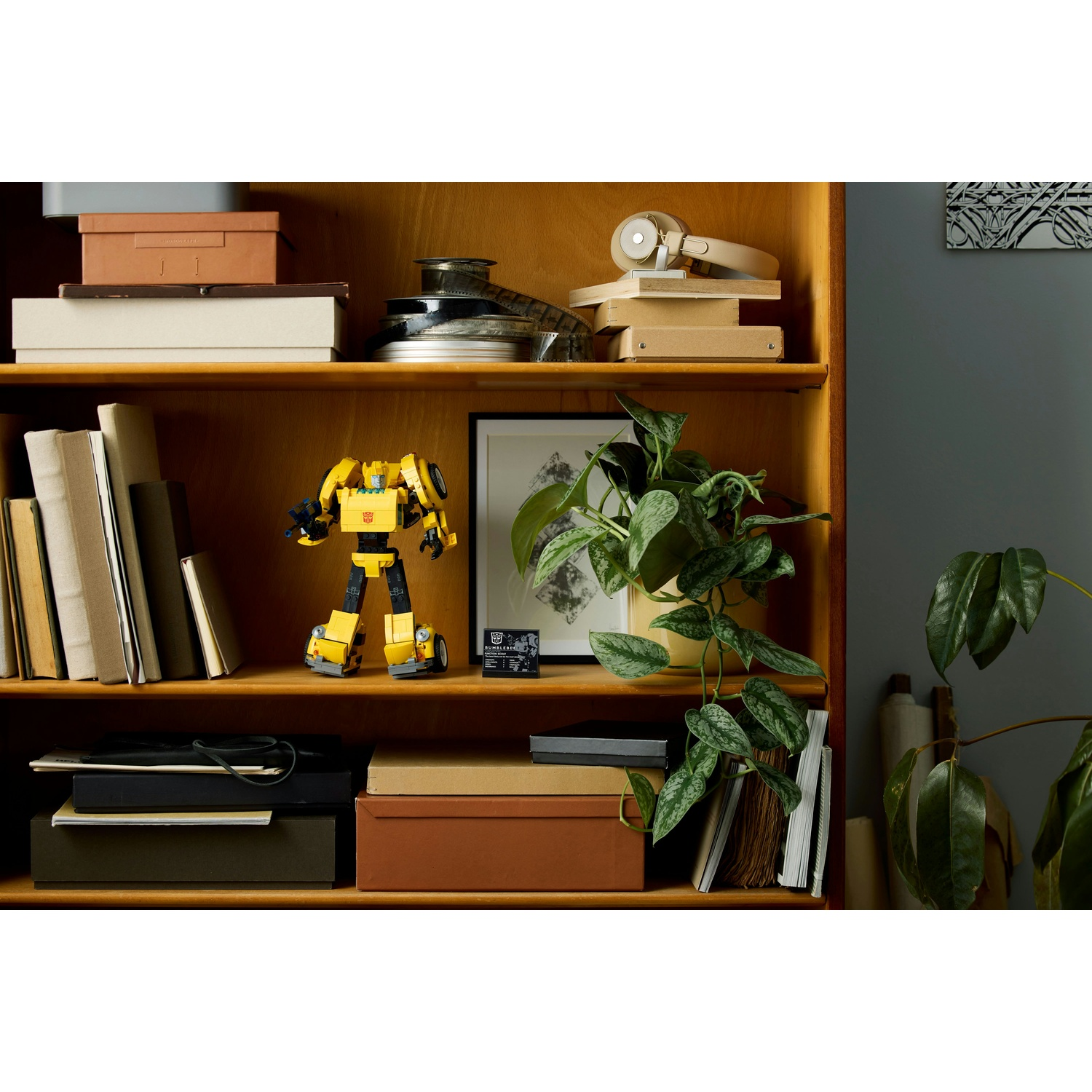 Una mensola di legno con oggetti. Un giocattolo robot giallo spicca. Sono presenti anche libri, scatole, una pianta e cuffie.