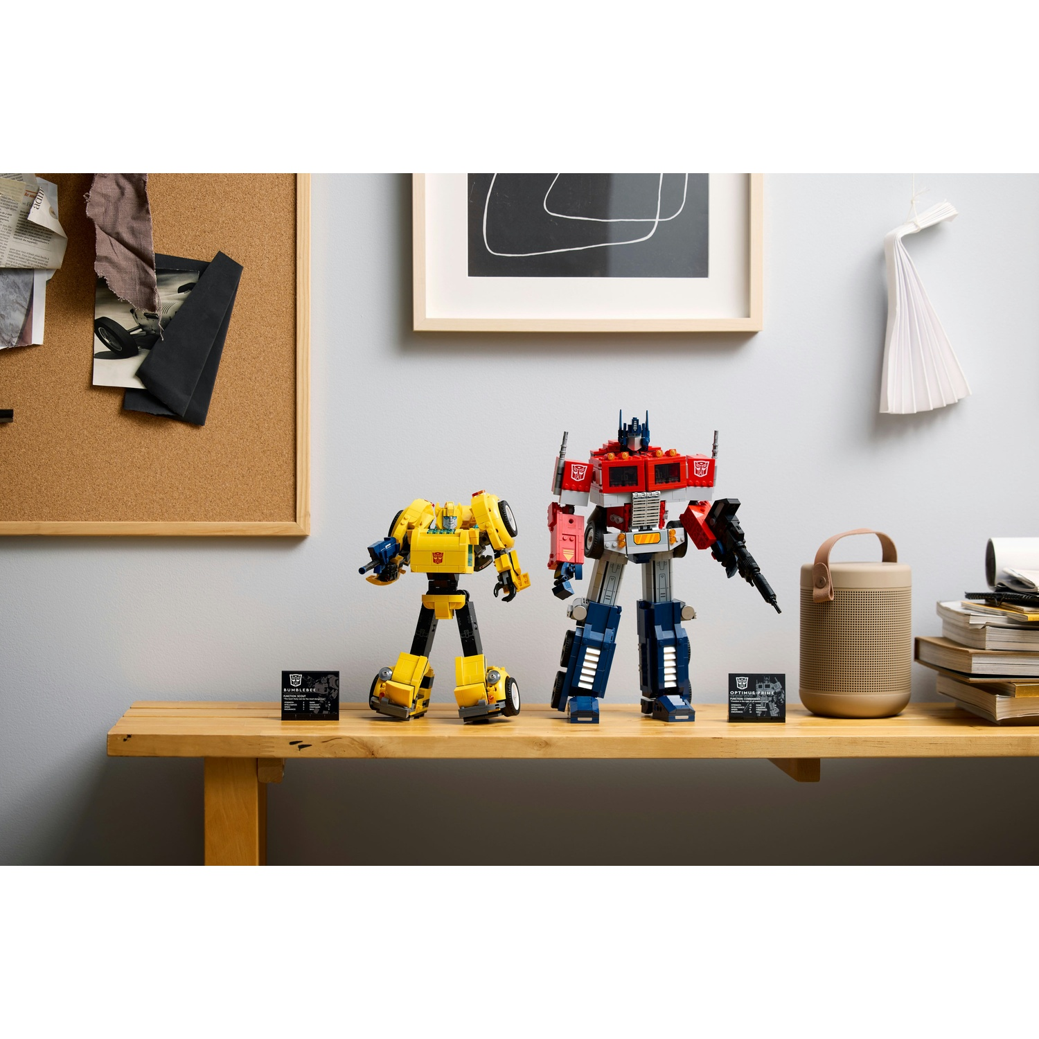 Due Transformers Lego, Bumblebee e Optimus Prime, su una mensola di legno. Un altoparlante e libri a destra. Bacheca e arte dietro.