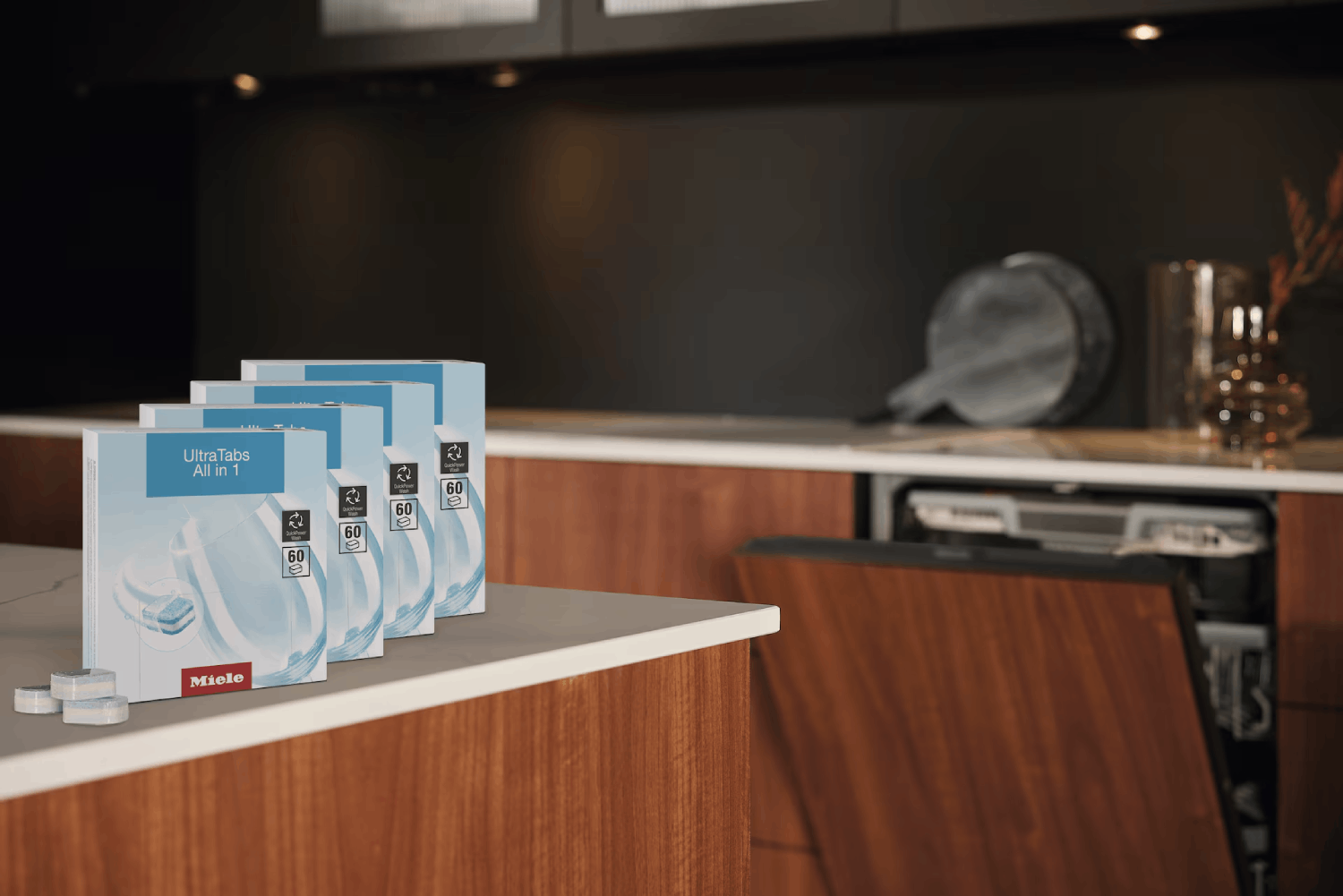 Cajas de detergente para lavavajillas con la etiqueta 'UltraTabs All in 1' en una encimera. Un lavavajillas está abierto en el fondo.