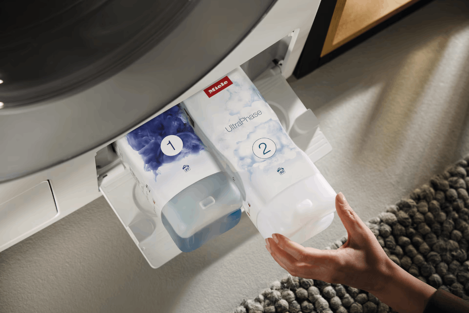 La mano de una persona está cerca de un cajón de lavadora con dos cartuchos de detergente etiquetados 1 y 2. La marca es Miele.