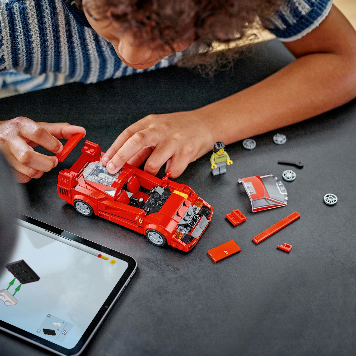 Un bambino assembla una Ferrari Lego rossa. I pezzi sono sparsi su una superficie nera. Un tablet mostra le istruzioni.