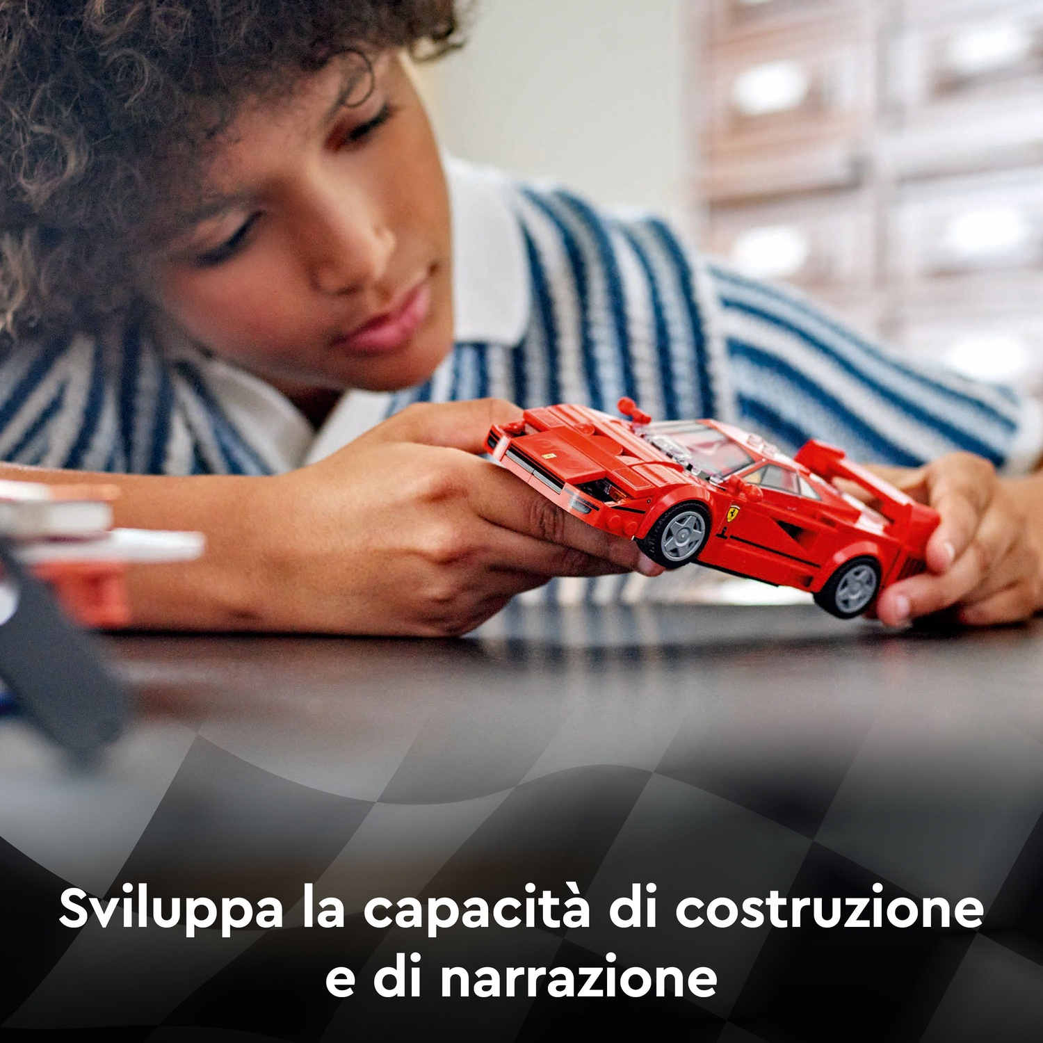 Un bambino tiene in mano un'auto giocattolo rossa. Sta guardando l'auto con attenzione. Lo sfondo è sfocato, compreso il testo.