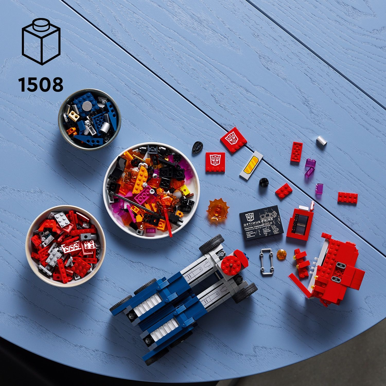 Componenti del set Lego su un tavolo blu. Include ciotole di mattoncini, un veicolo blu e bianco e pezzi Lego rossi e neri.