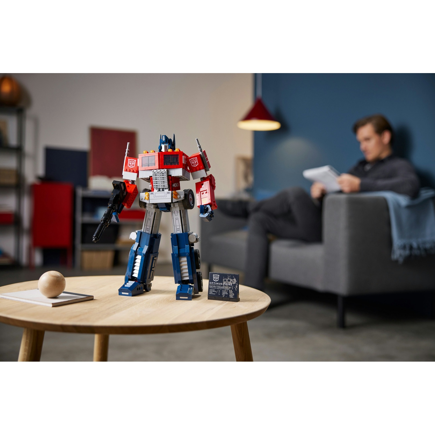 Un Optimus Prime Lego è su un tavolo. Un uomo legge un libro su un divano sullo sfondo. Una palla di legno e una carta sono sul tavolo.