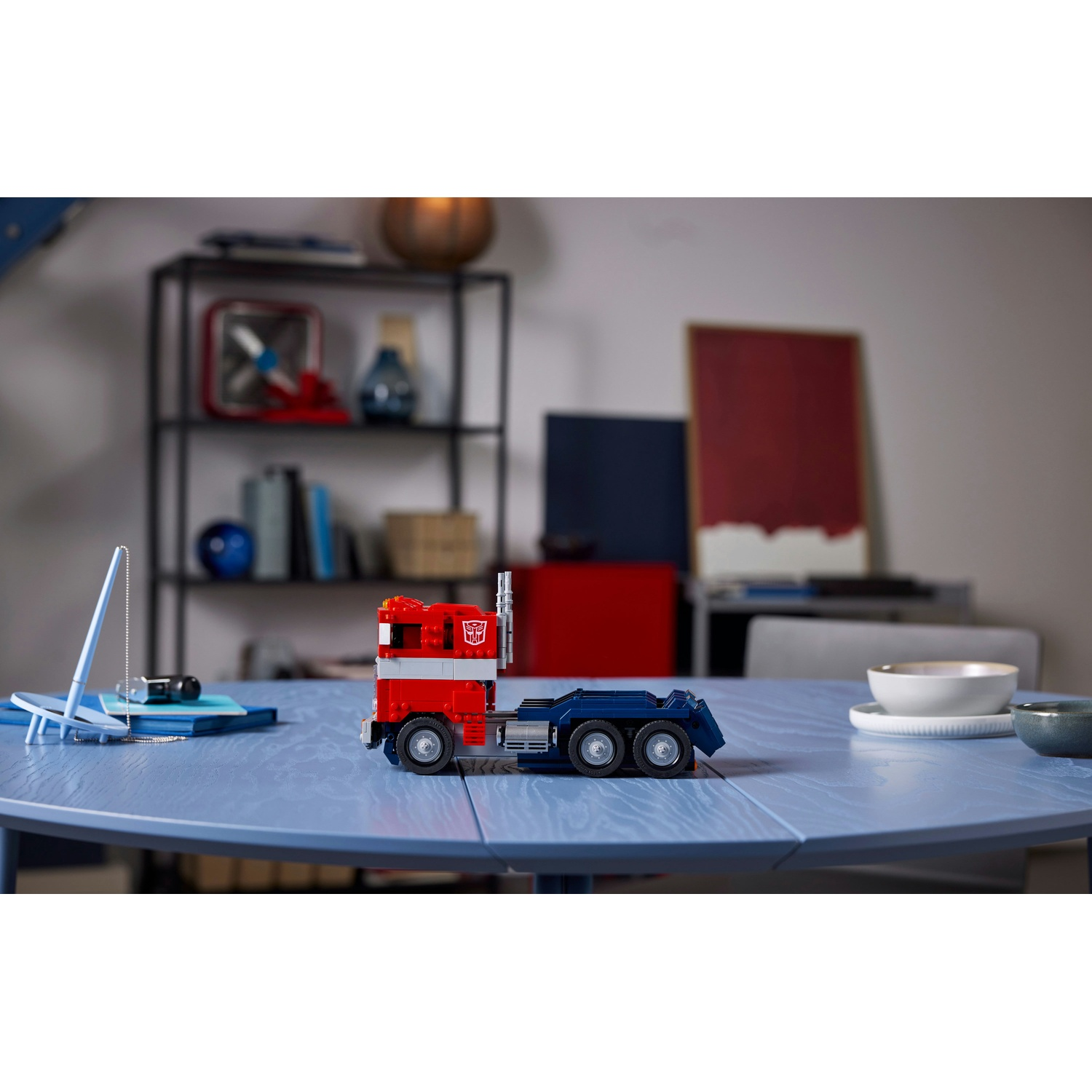 Un camion Lego Optimus Prime è su un tavolo blu. Lo sfondo include una mensola, un muro con dipinti e una ciotola.