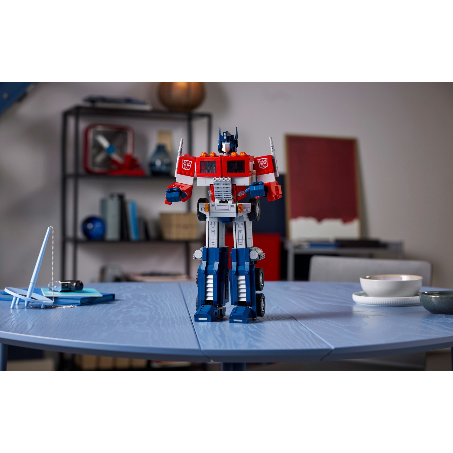 Un Lego Optimus Prime è su un tavolo blu. Lo sfondo mostra scaffali, un dipinto e una ciotola. Il giocattolo è rosso, blu e argento.