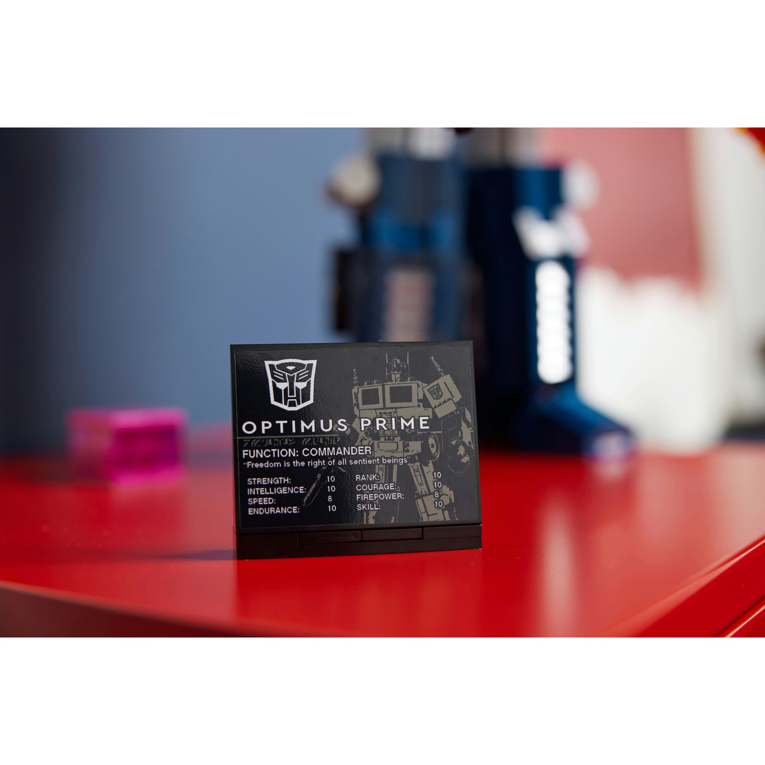 Una carta nera con il logo e le statistiche di Optimus Prime è su una superficie rossa. Un cubo rosa e un oggetto blu sullo sfondo.