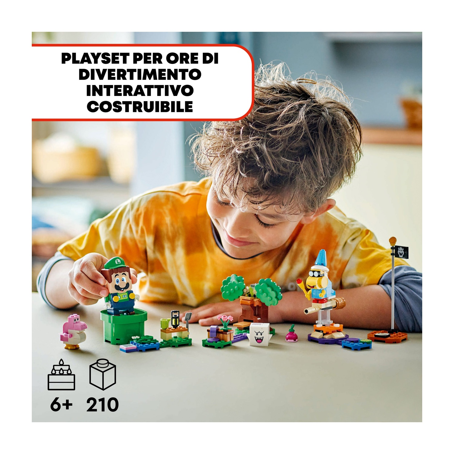 Un ragazzino gioca con un set Lego con Luigi, un dinosauro rosa e altri personaggi su un tavolo, sorridendo.