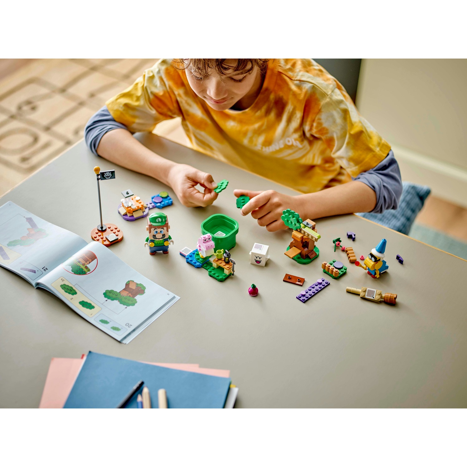 Un ragazzo costruisce un set Lego con i personaggi di Mario. Il set include Luigi, un maiale e altri elementi su un tavolo grigio.
