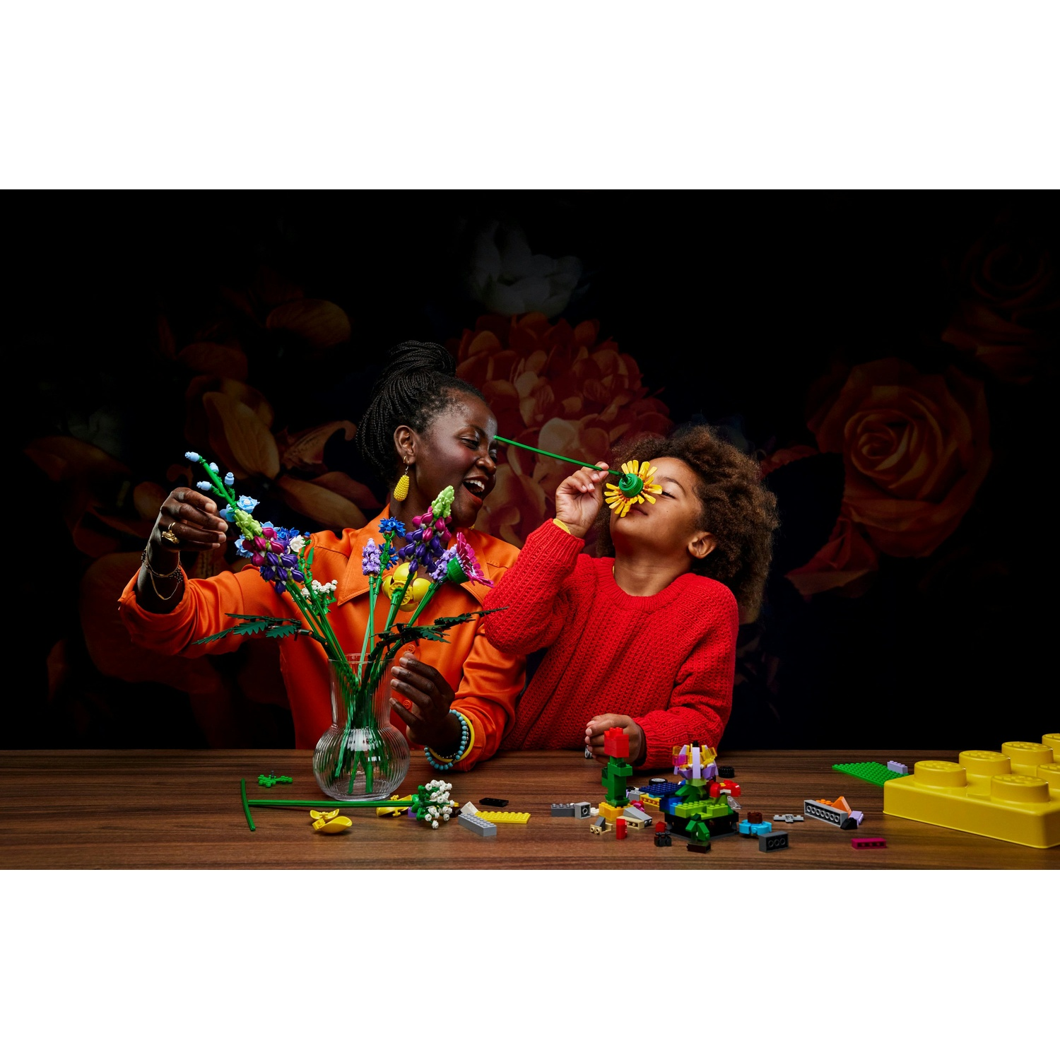 Una donna e un bambino giocano con fiori Lego. Il bambino si avvicina un girasole al naso. La donna sorride.