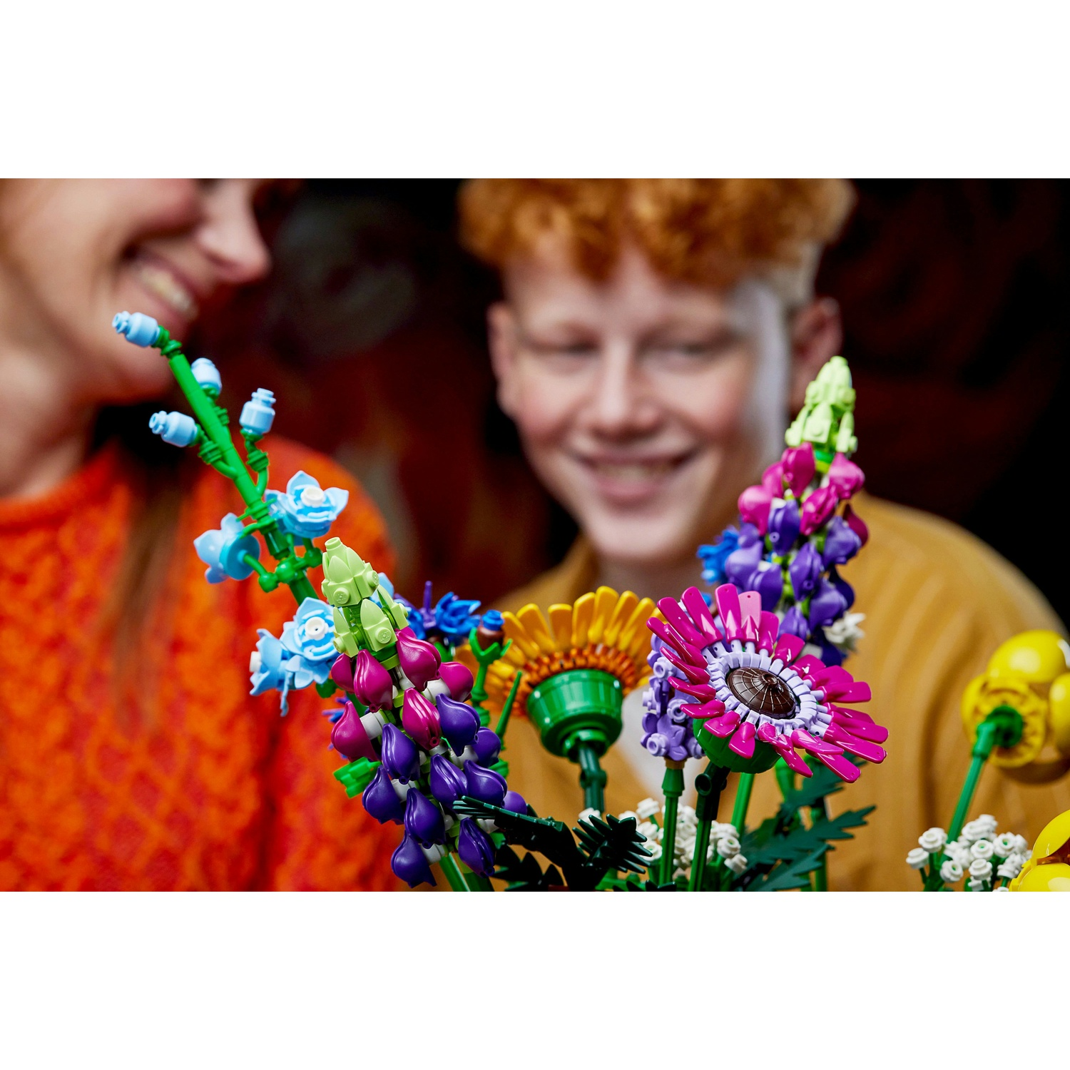 Fiori Lego colorati in un bouquet, con due persone sorridenti sfocate sullo sfondo. Colori vivaci come rosa, blu e giallo.