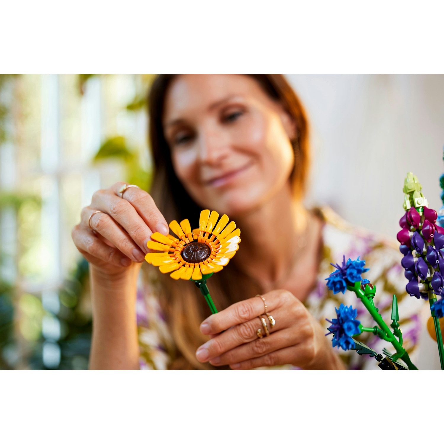 Una donna tiene in mano un fiore Lego giallo. Altri fiori Lego blu, viola e verdi sullo sfondo. La donna sorride.