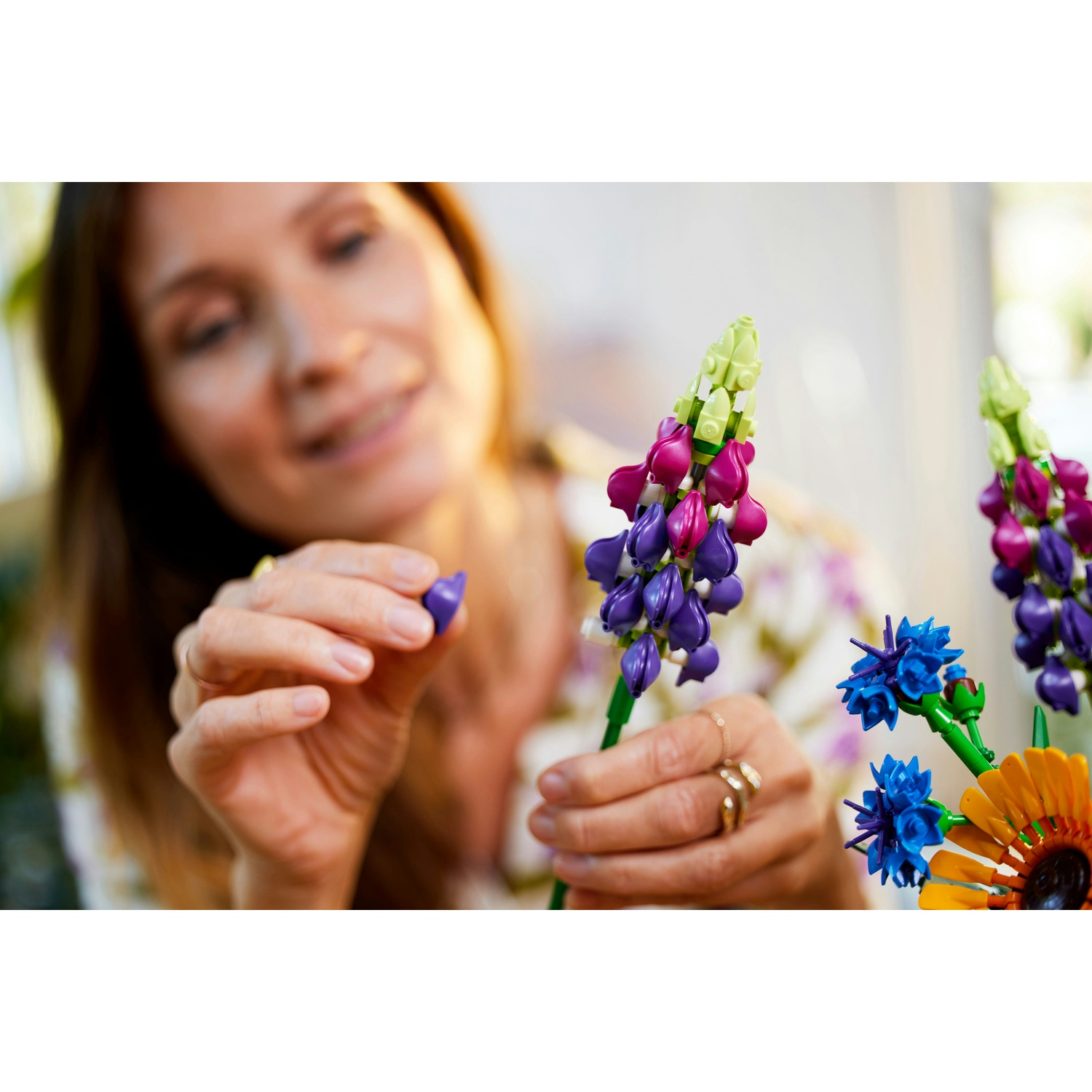 Una donna assembla fiori Lego colorati. Fiori viola, rosa e blu sono a fuoco. La donna indossa una camicia floreale.