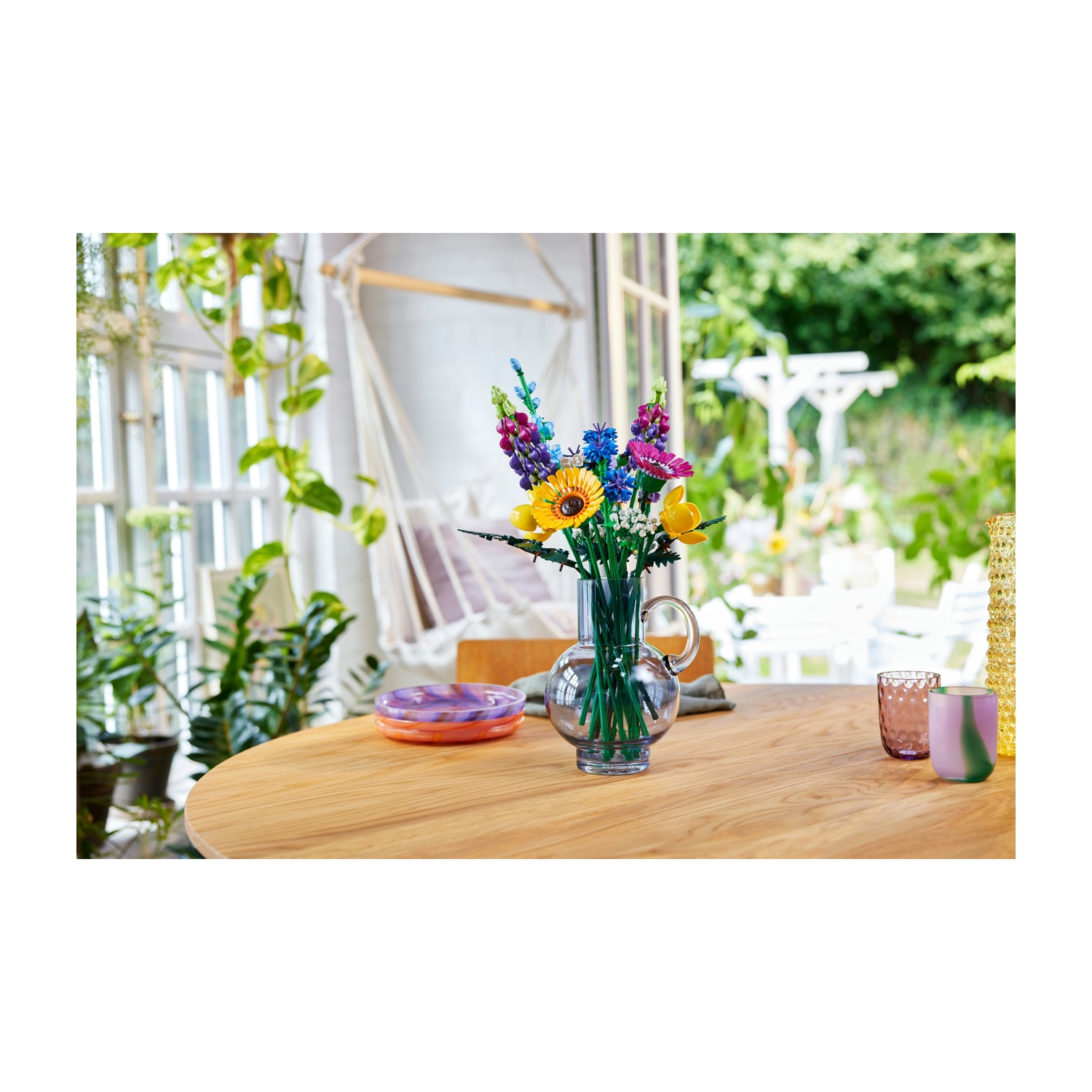 Un tavolo di legno con un vaso di vetro con fiori colorati. Due bicchieri e un piatto sul tavolo. Fogliame verde sullo sfondo.