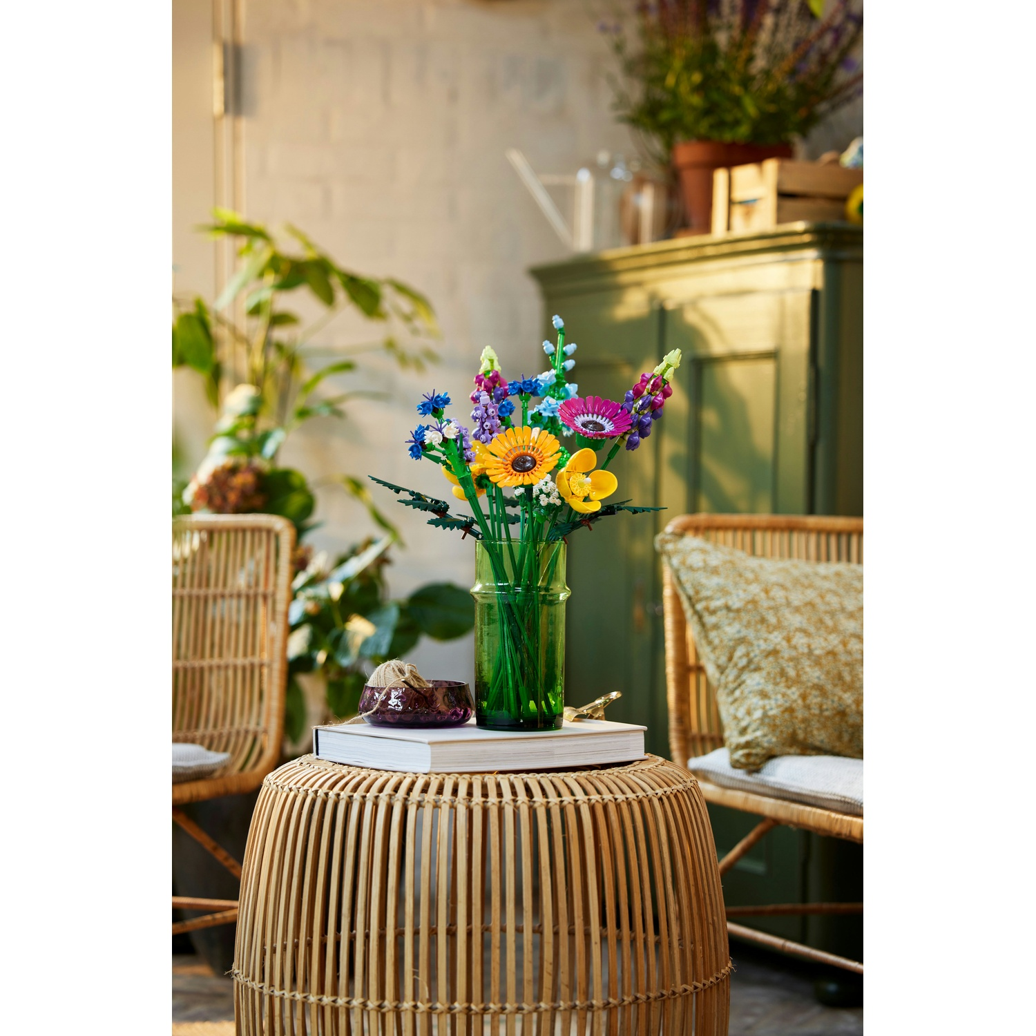 Fiori colorati in un vaso verde su un tavolo di vimini. Un libro e una ciotola decorativa sono anche sul tavolo. Ci sono sedie nelle vicinanze.