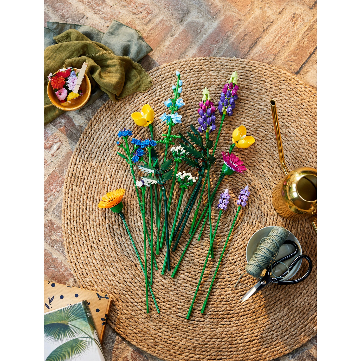 Fiori artificiali colorati su un tappeto intrecciato. Include un annaffiatoio, forbici e una ciotola con spago e altri fiori.